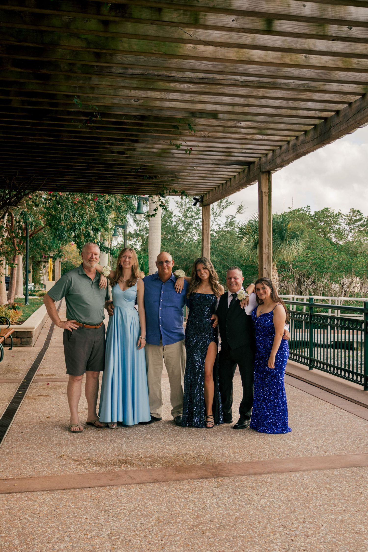 Group of six people posing in formal attire under a wooden pergola at an outdoor venue.