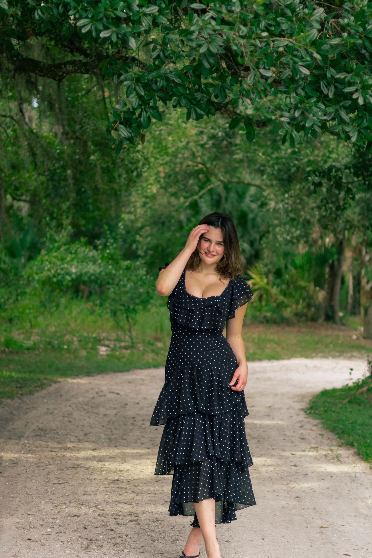 Smiling woman in black polka dot dress walking on garden path under canopy of green trees.