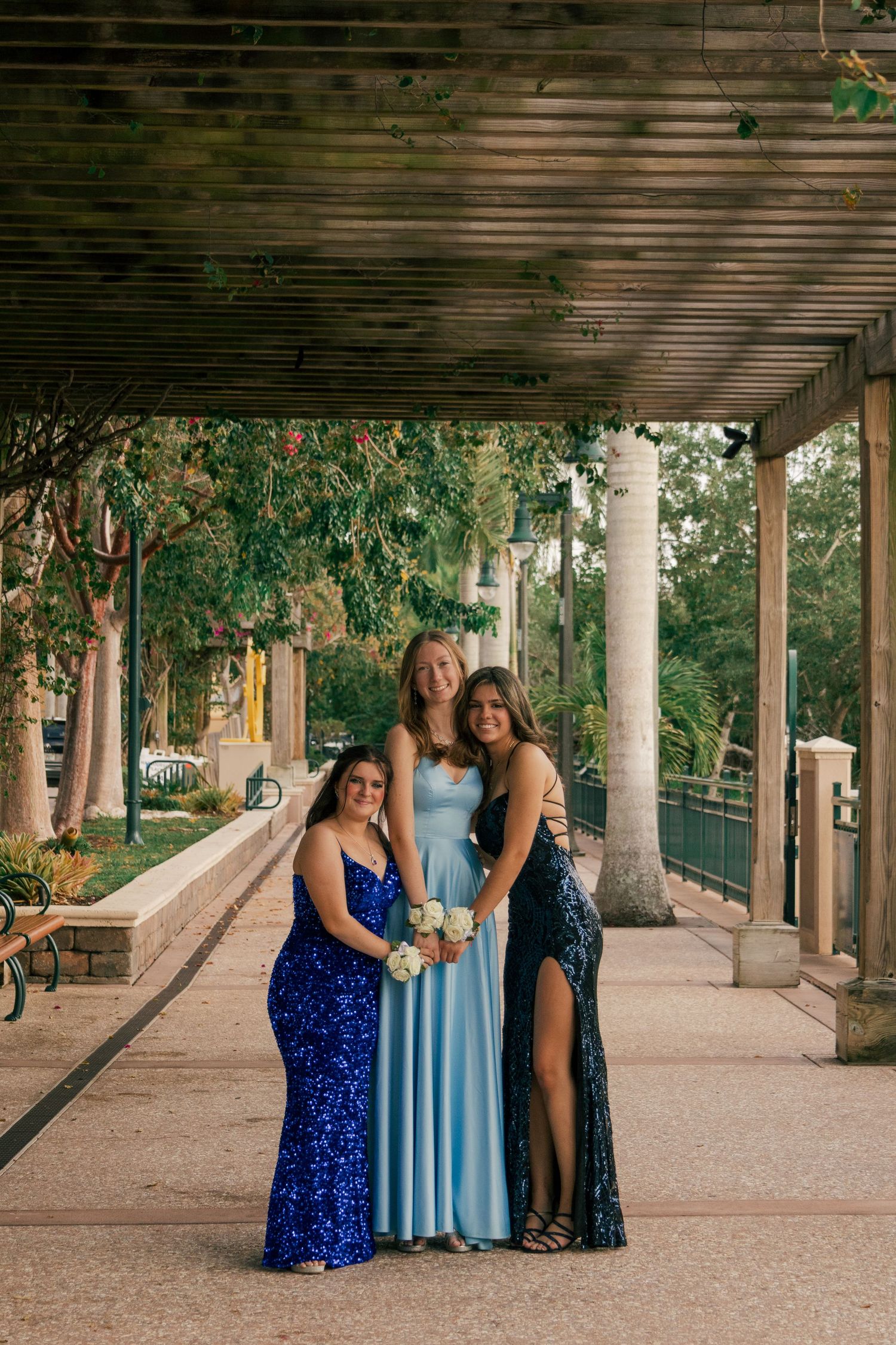 Three young women in blue prom dresses posing under a pergola in an outdoor walkway.