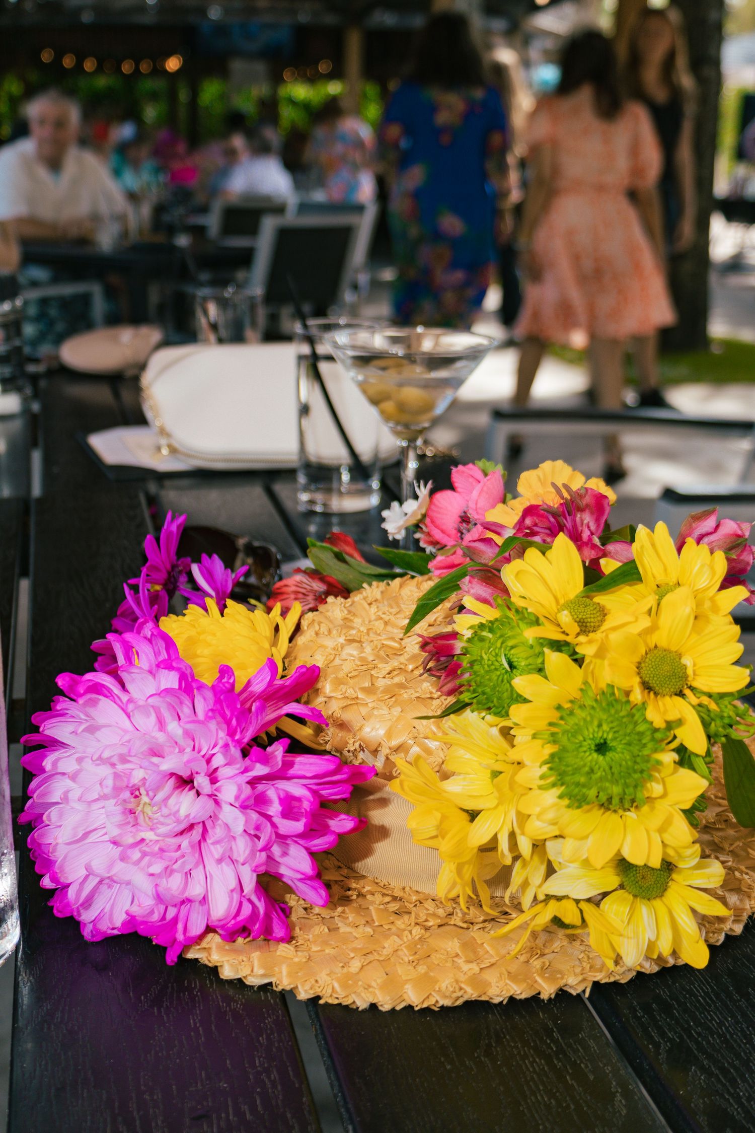 Colorful bouquet of pink and yellow flowers in a straw hat on a dark wooden table at an outdoor event.