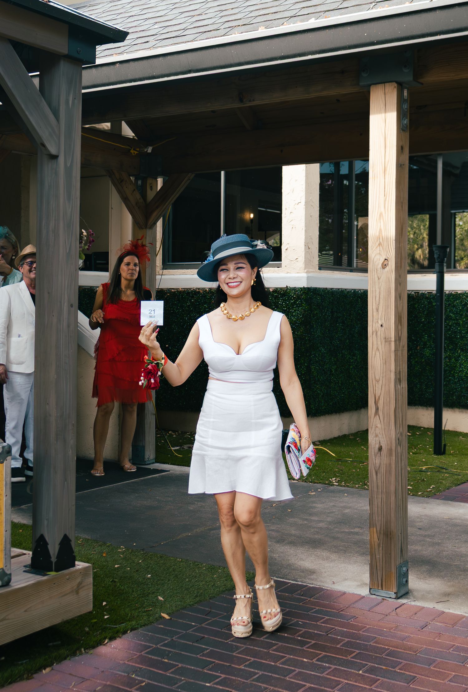 Woman in white dress and blue hat smiles walking under a gazebo at an outdoor social event.