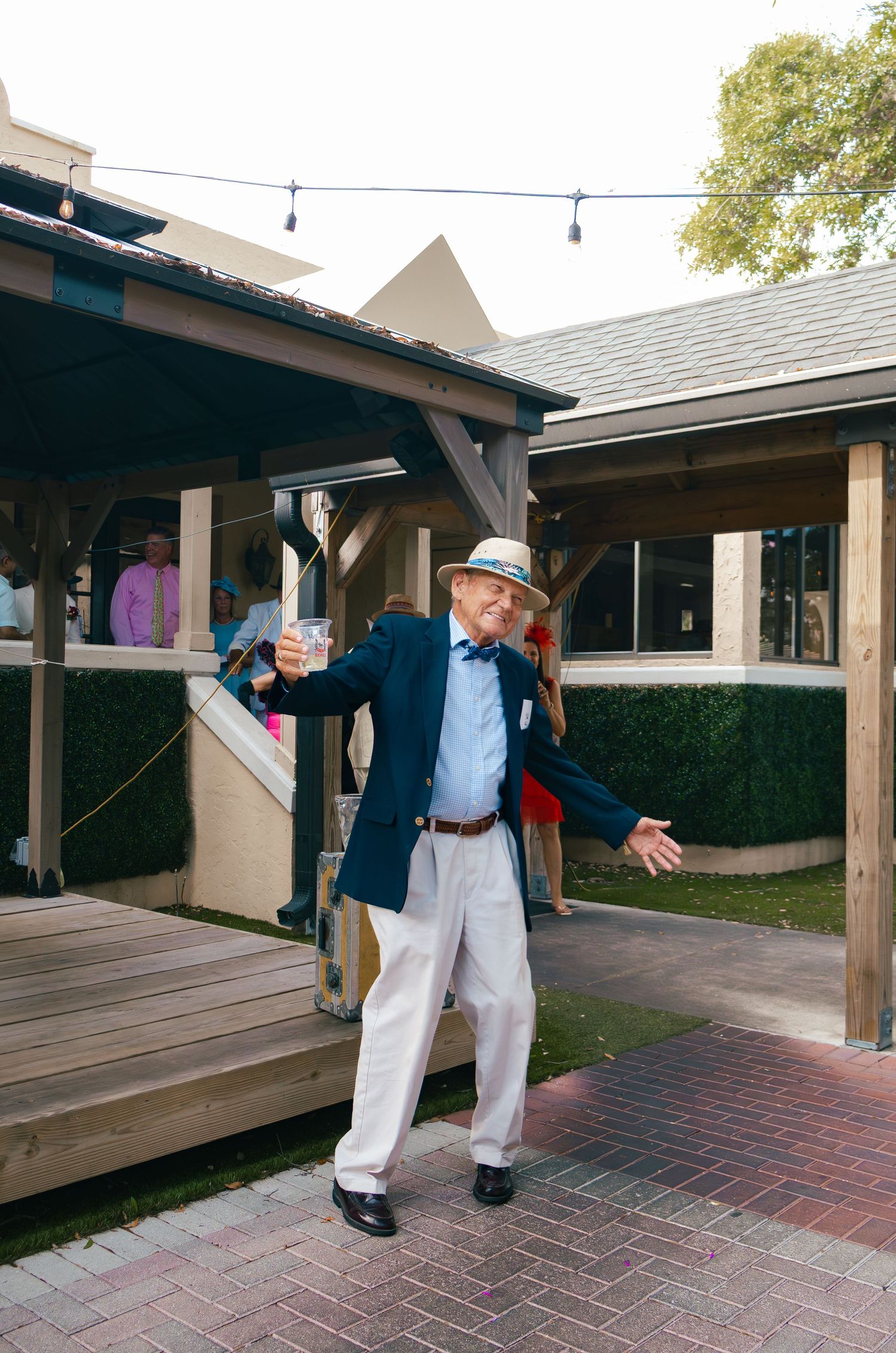 Elderly man in navy blazer, bow tie, and straw hat raises a glass outdoors at an event near a wooden gazebo.
