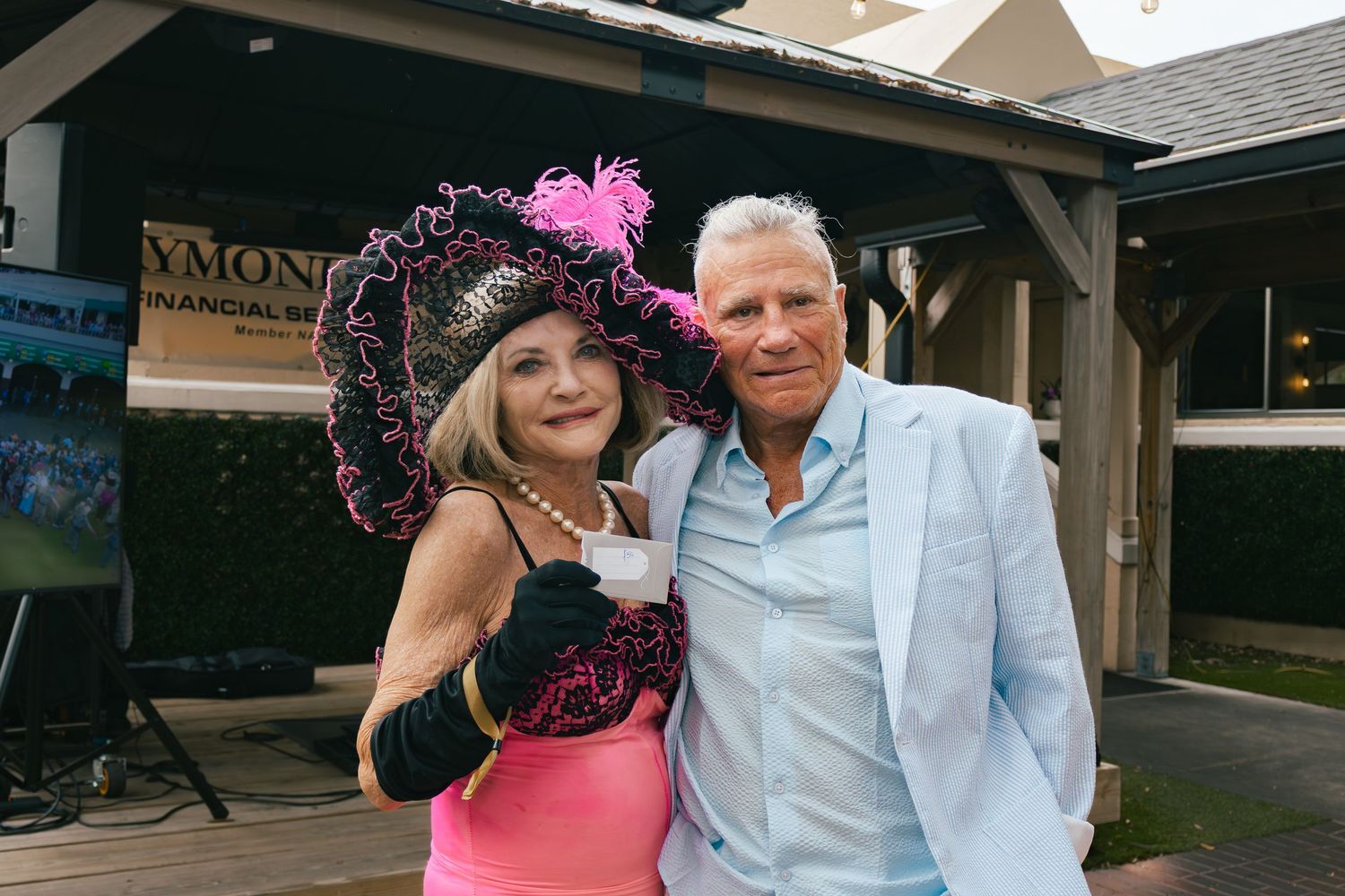 Woman in pink dress and elaborate black feathered hat poses with man in light blue blazer.