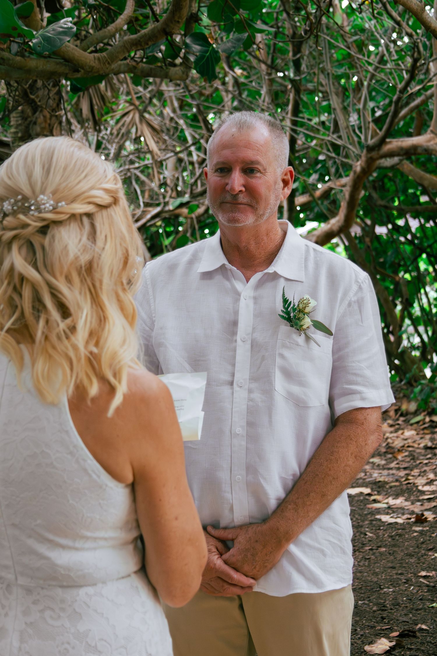 Groom with boutonniere holding hands with blonde bride while exchanging vows at an outdoor tropical wedding ceremony.