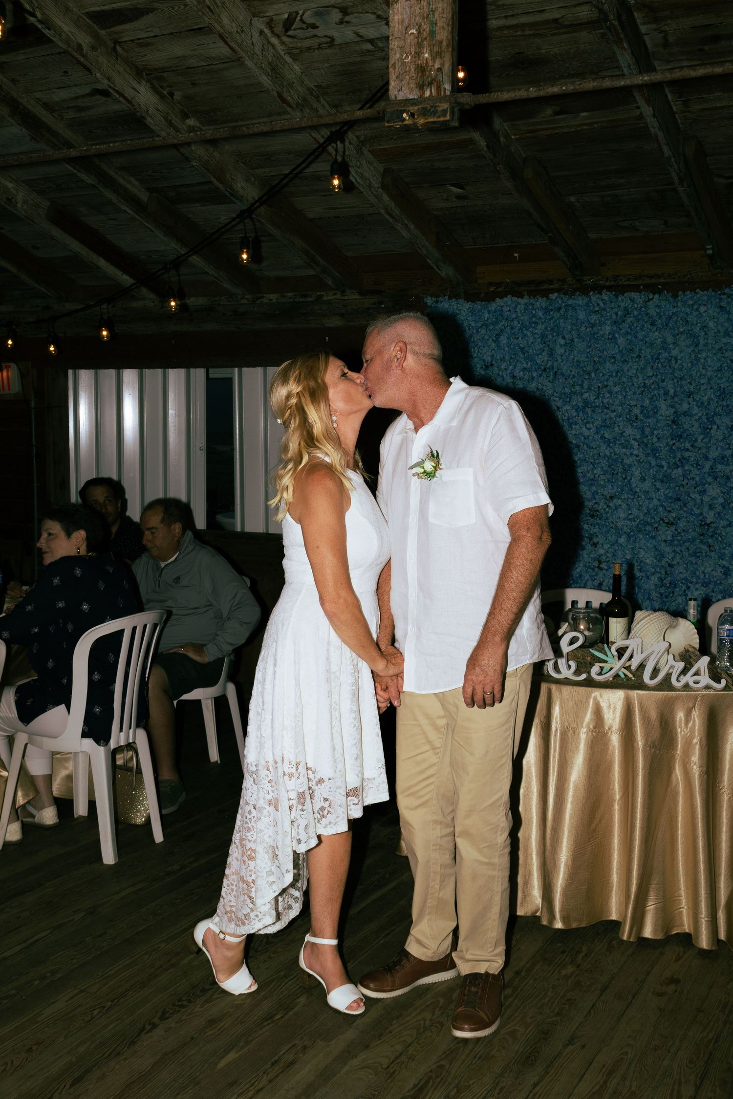 Bride and groom share a romantic kiss while holding hands at their rustic wedding reception.