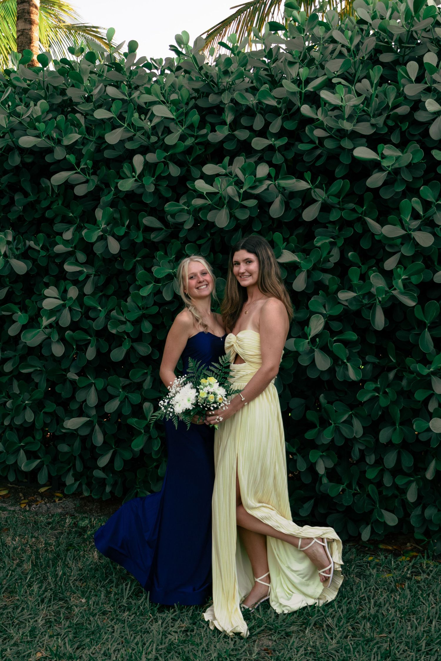 Two young women in navy and pale yellow formal gowns smile together before green hedge.