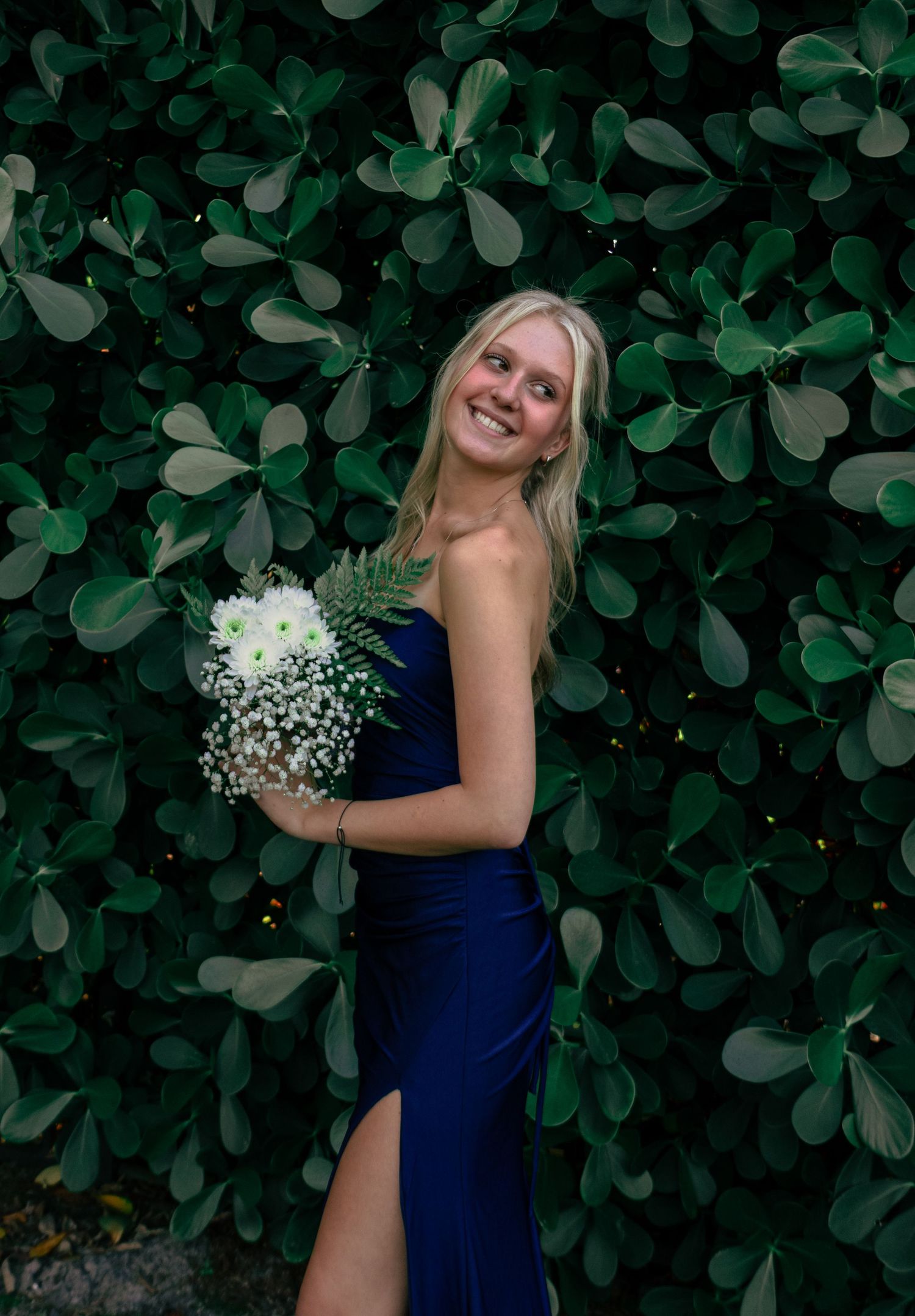 Smiling blonde teen in a navy strapless gown holding a white floral bouquet at prom.