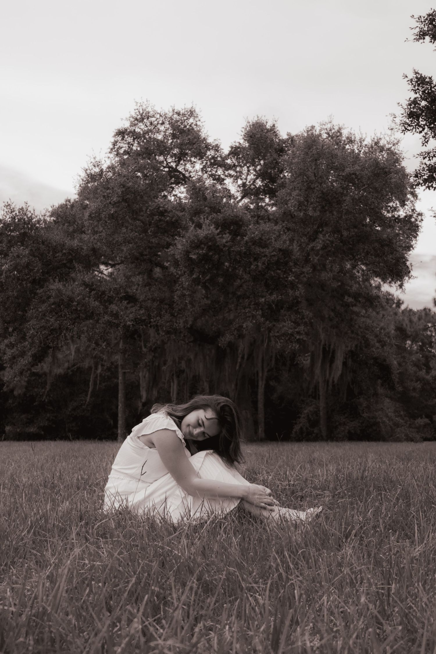 Black and white photo of young woman in white dress sitting alone in open field near trees.