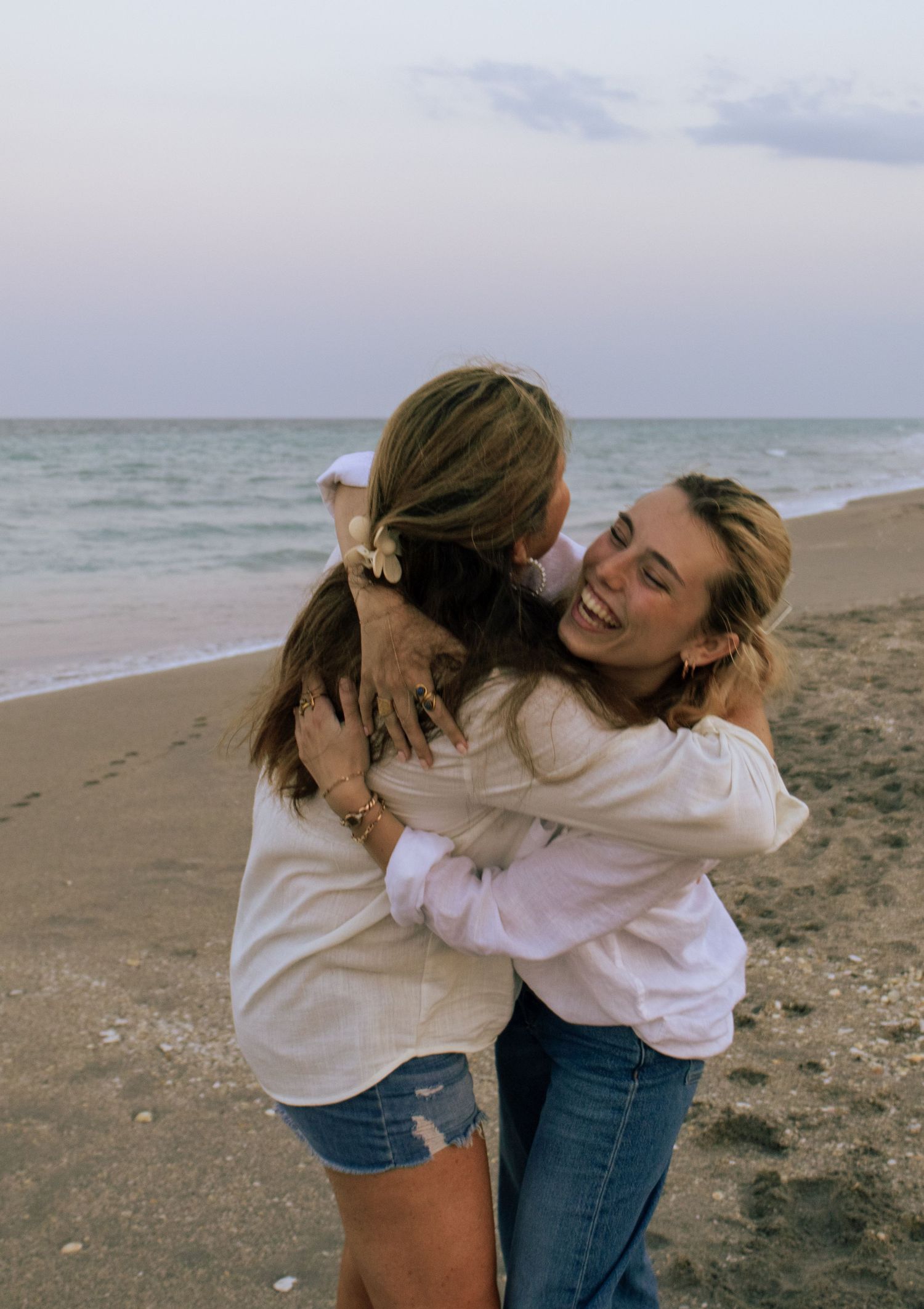 Two young women hugging and laughing on a sandy beach at dusk wearing white shirts.