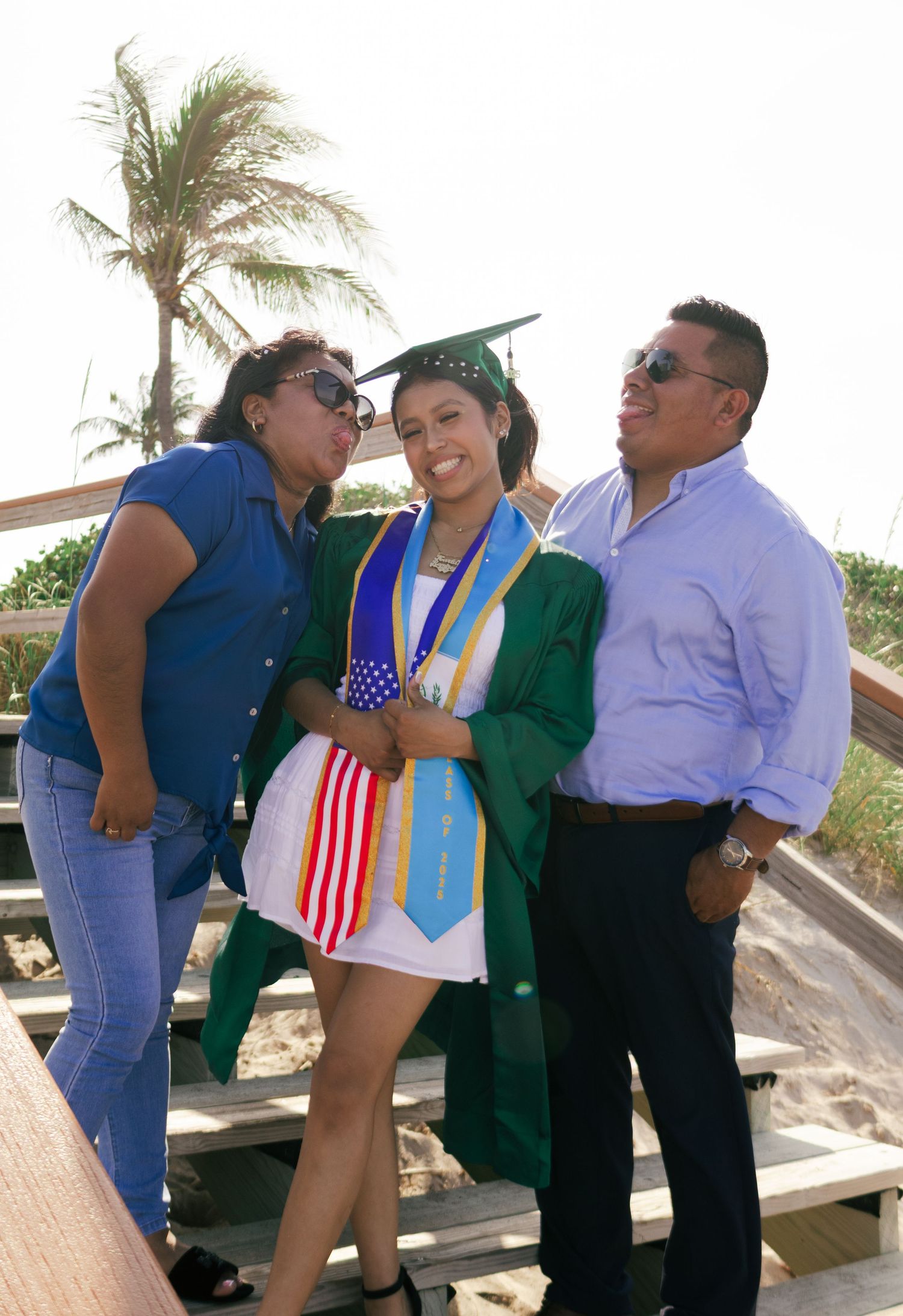 Graduate in green cap and gown with American flag stole celebrates with proud parents at a beach setting.