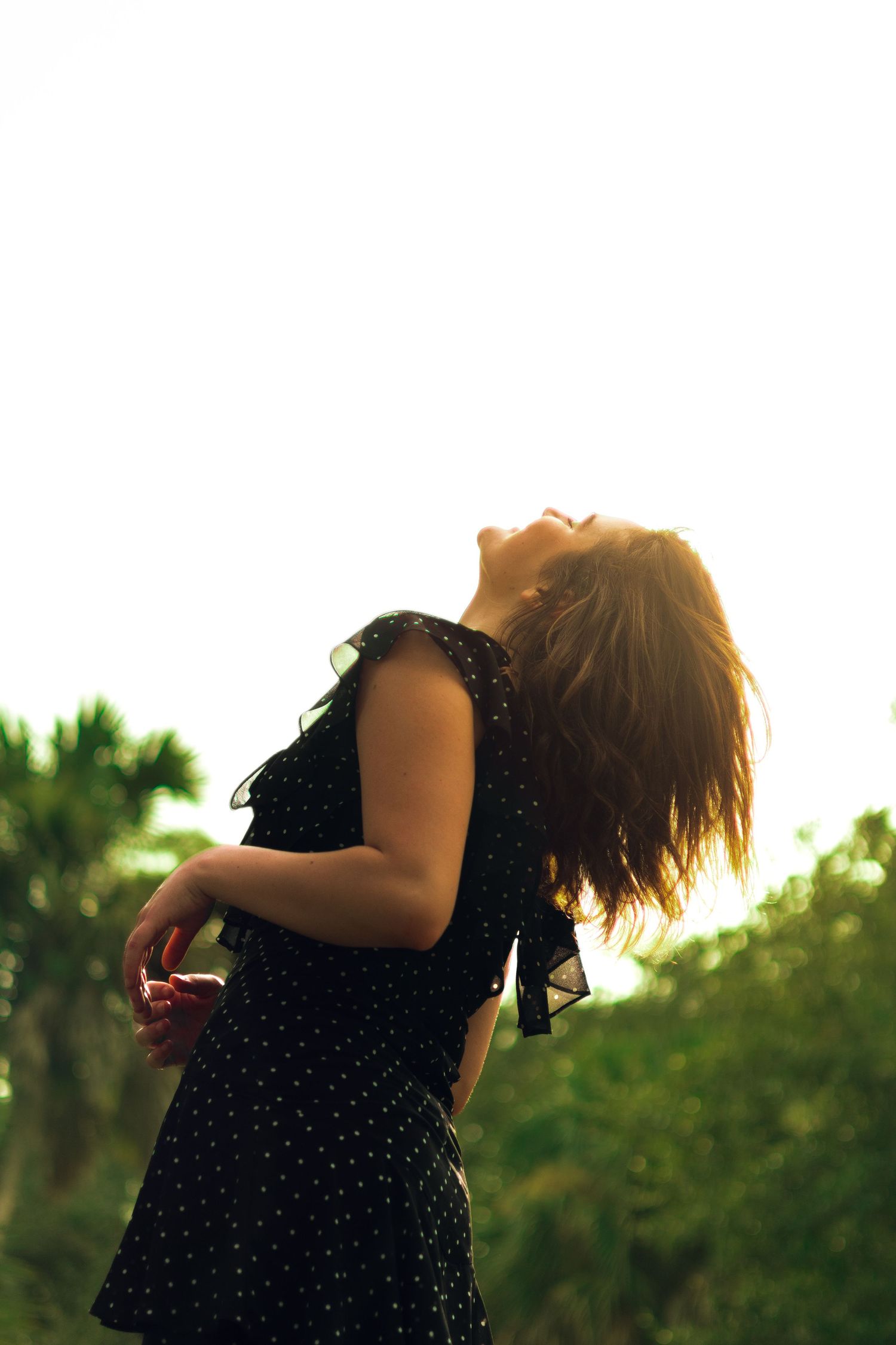 Woman in black polka dot dress tossing hair in golden backlit sunlight outdoors.