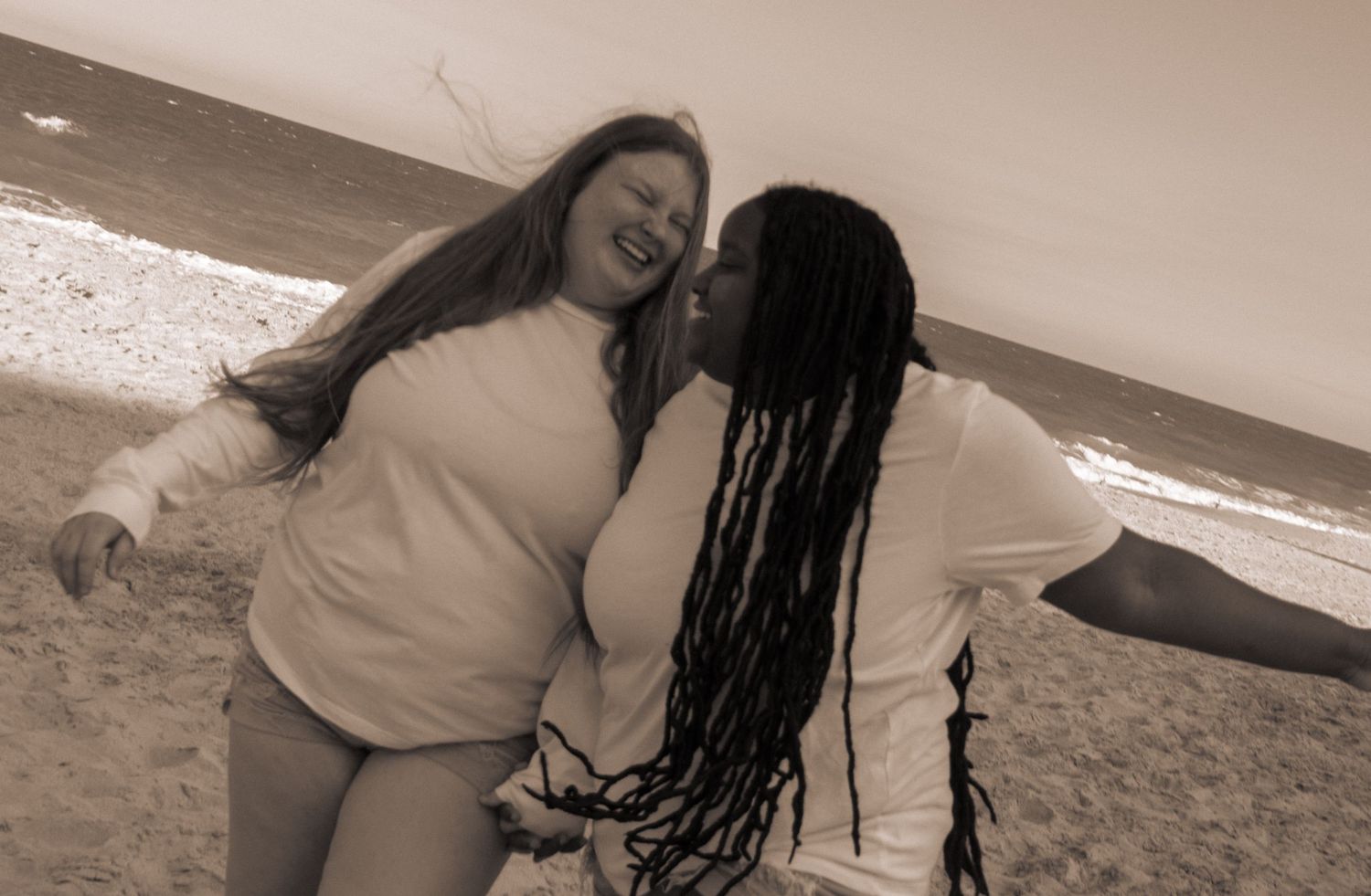 Two women laughing and embracing on a windy beach in sepia tone.