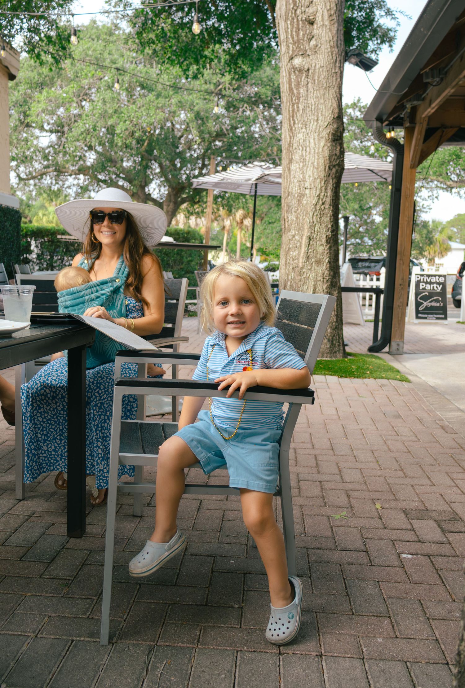 Young blonde toddler sits in patio chair while mother in wide-brim hat reviews menu at outdoor restaurant.