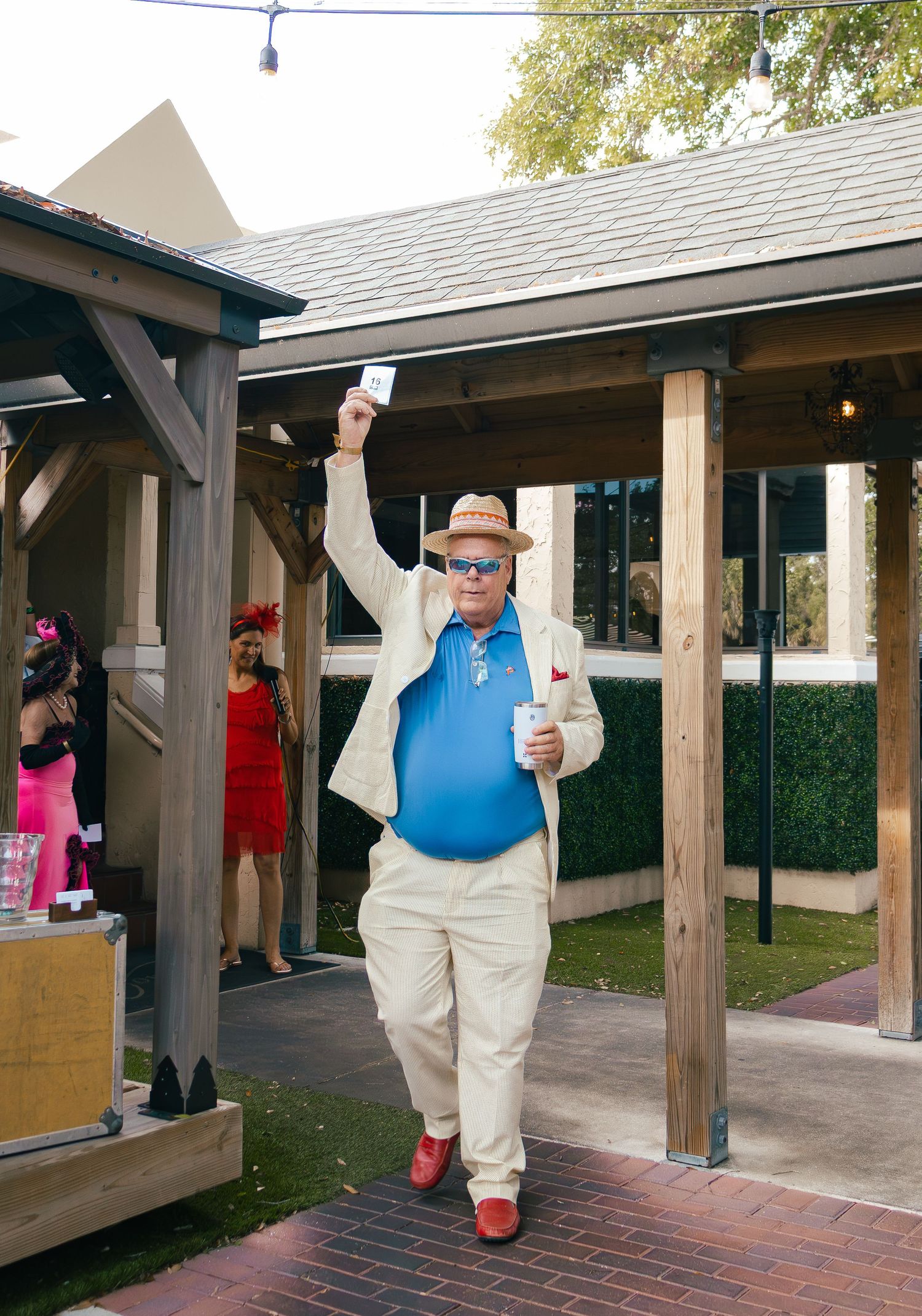 Man in cream suit, blue shirt, and red shoes raises card while holding drink at outdoor event.