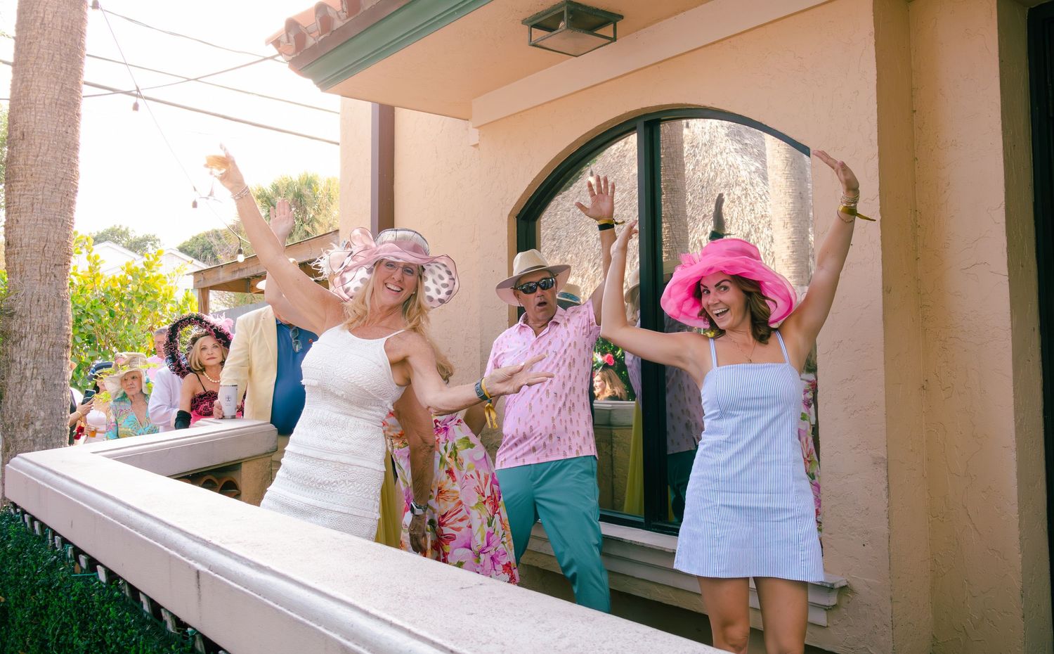 Group of women in festive hats celebrate with raised arms at outdoor party.