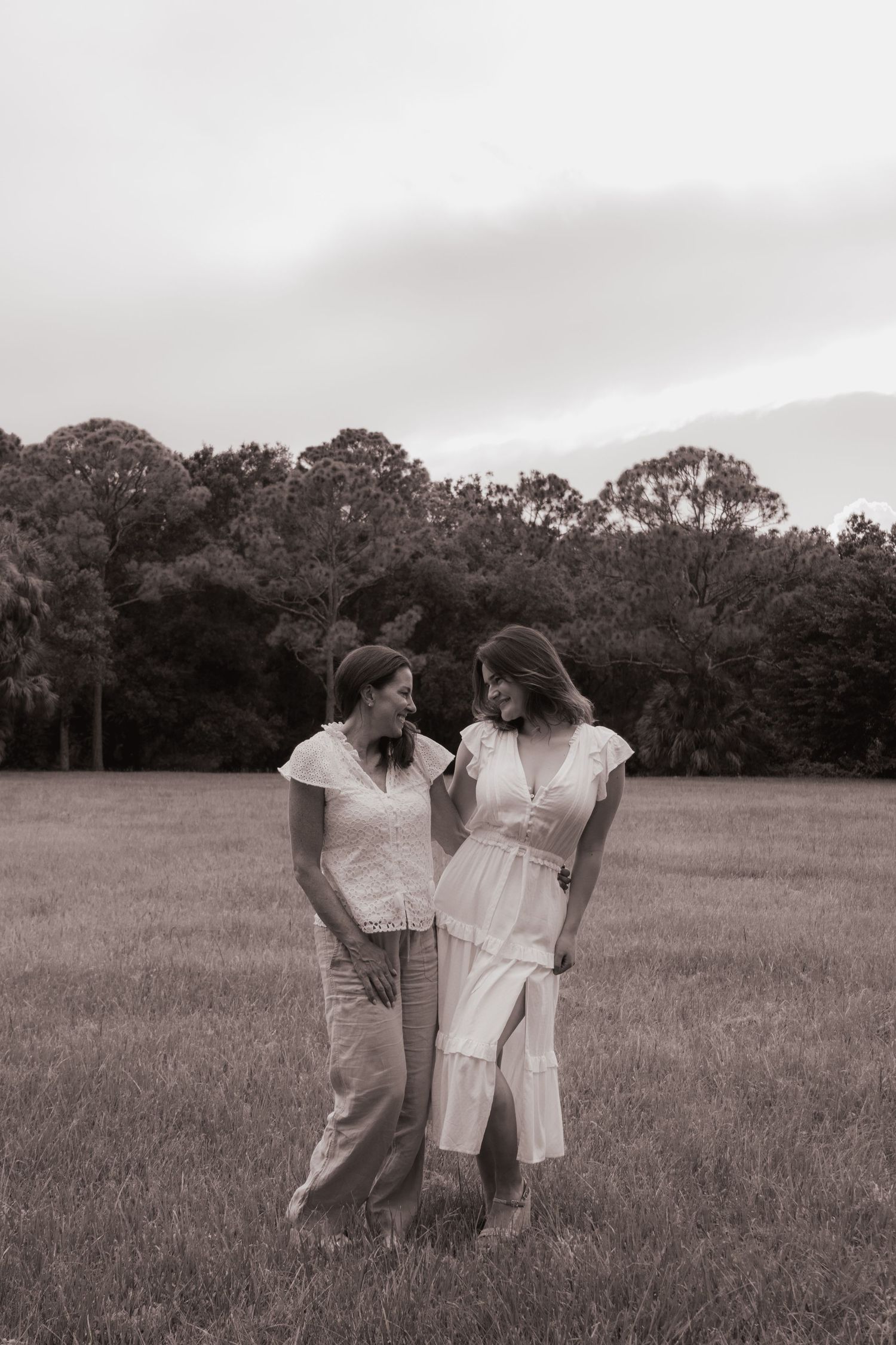 Black and white photo of two women standing together in open field, smiling at each other.