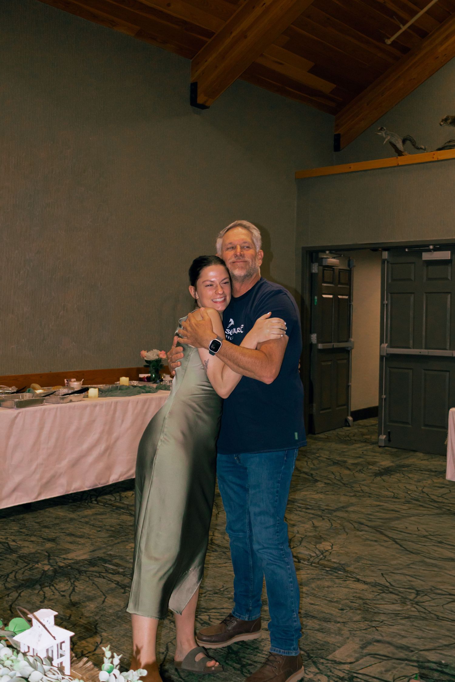 Woman in sage green dress hugs older man in navy shirt at wedding reception venue.