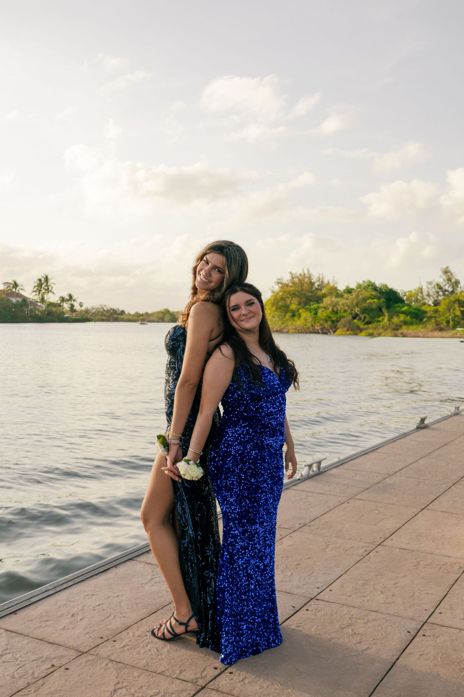 Two smiling girls in blue sequin formal dresses pose back-to-back on a scenic waterfront dock.