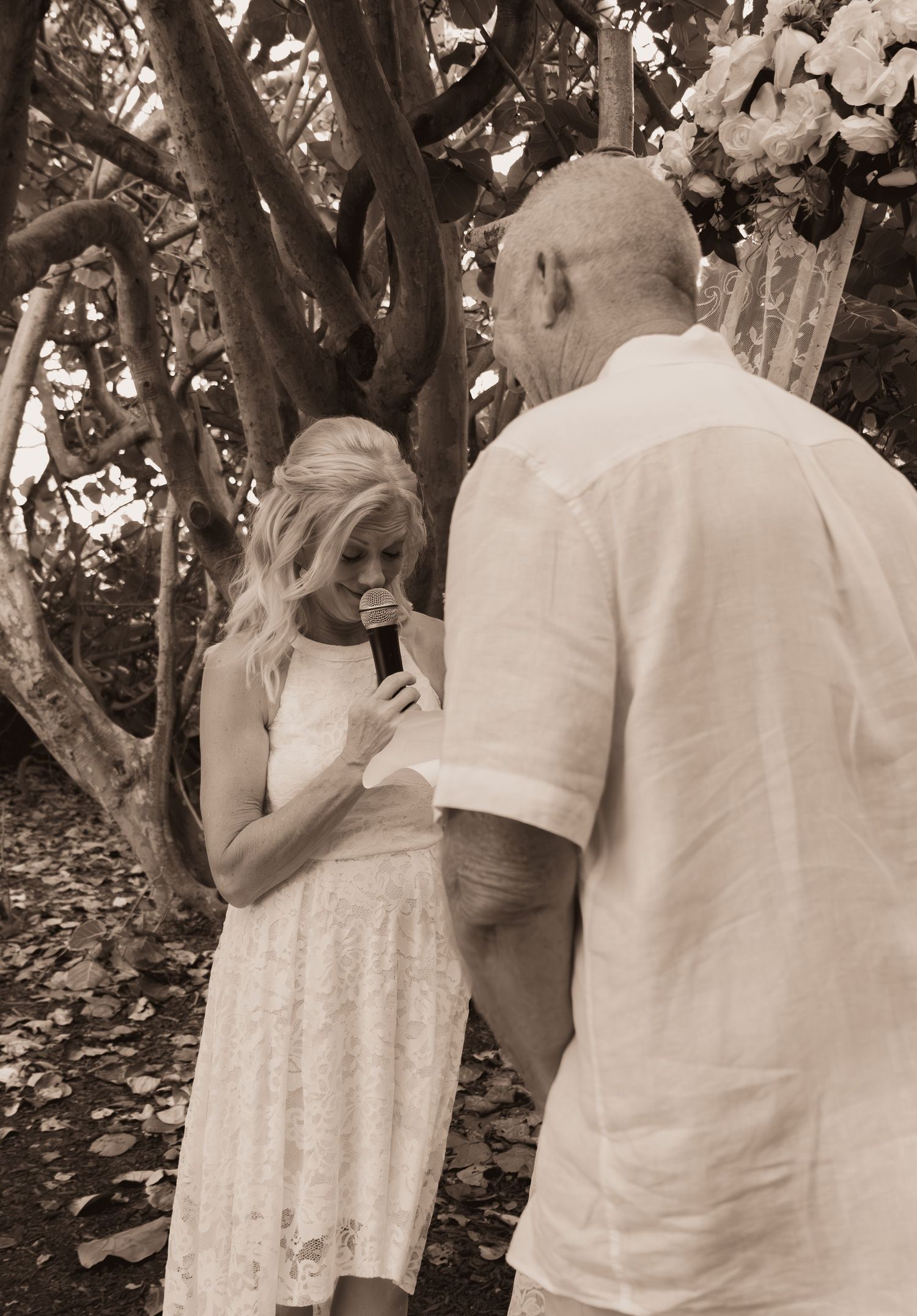 Sepia-toned image of a bride emotionally reading her wedding vows into a microphone during an outdoor ceremony.