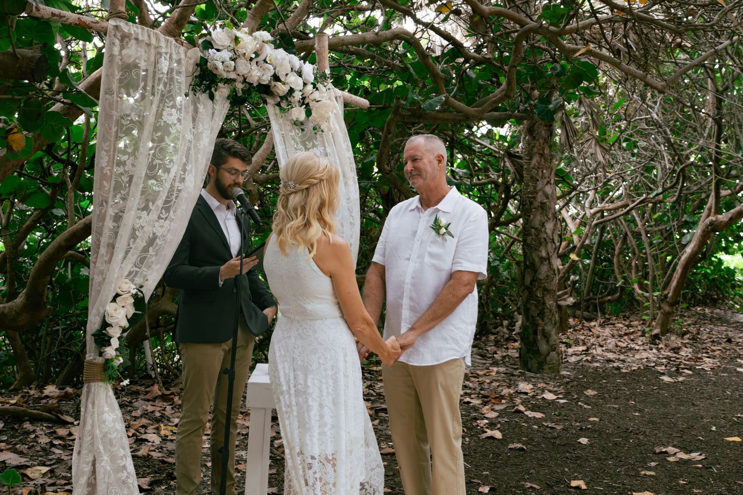Couple exchanges vows under floral arch in tropical outdoor wedding ceremony.