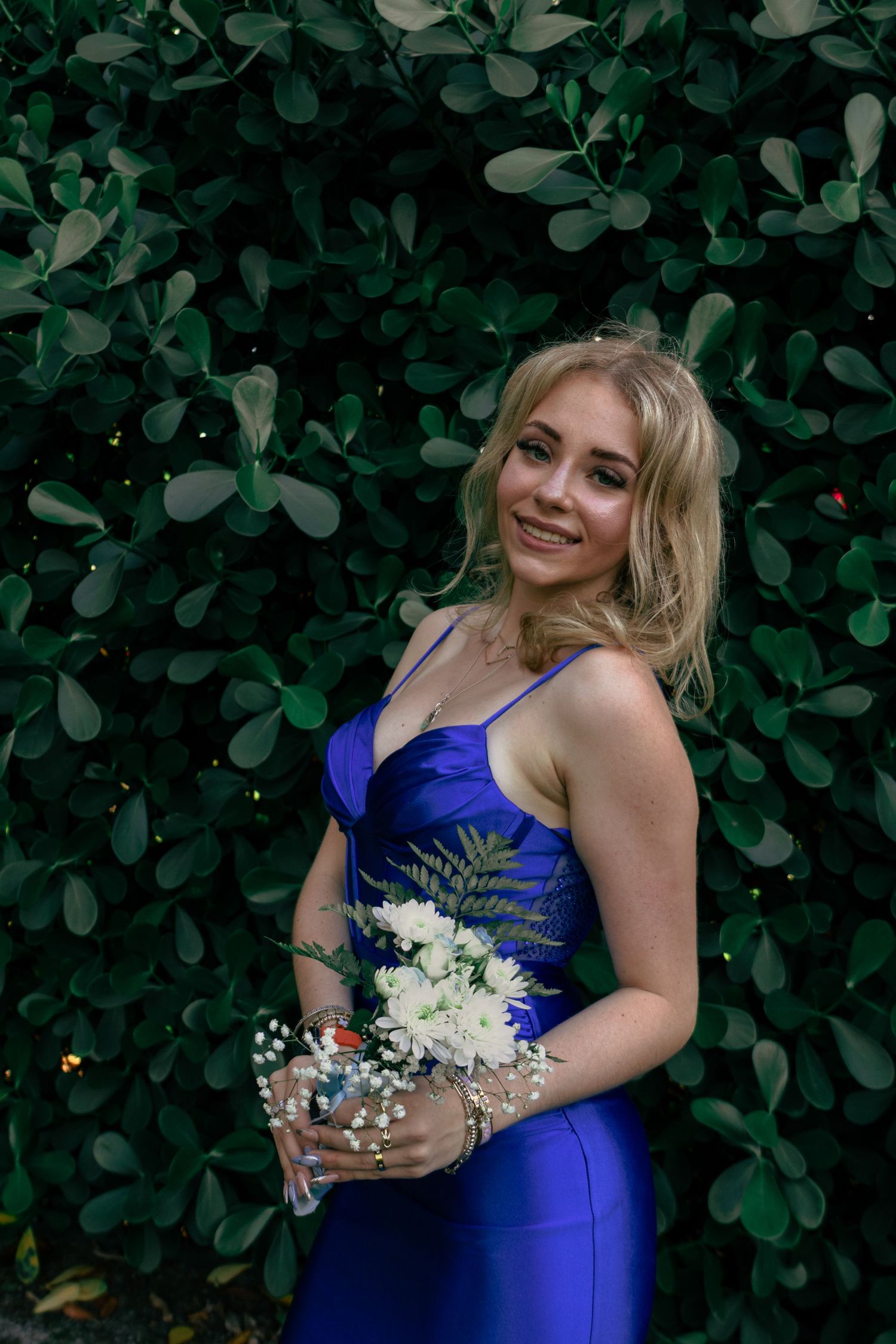 Smiling blonde in a royal blue spaghetti strap prom dress holding a white flower bouquet.