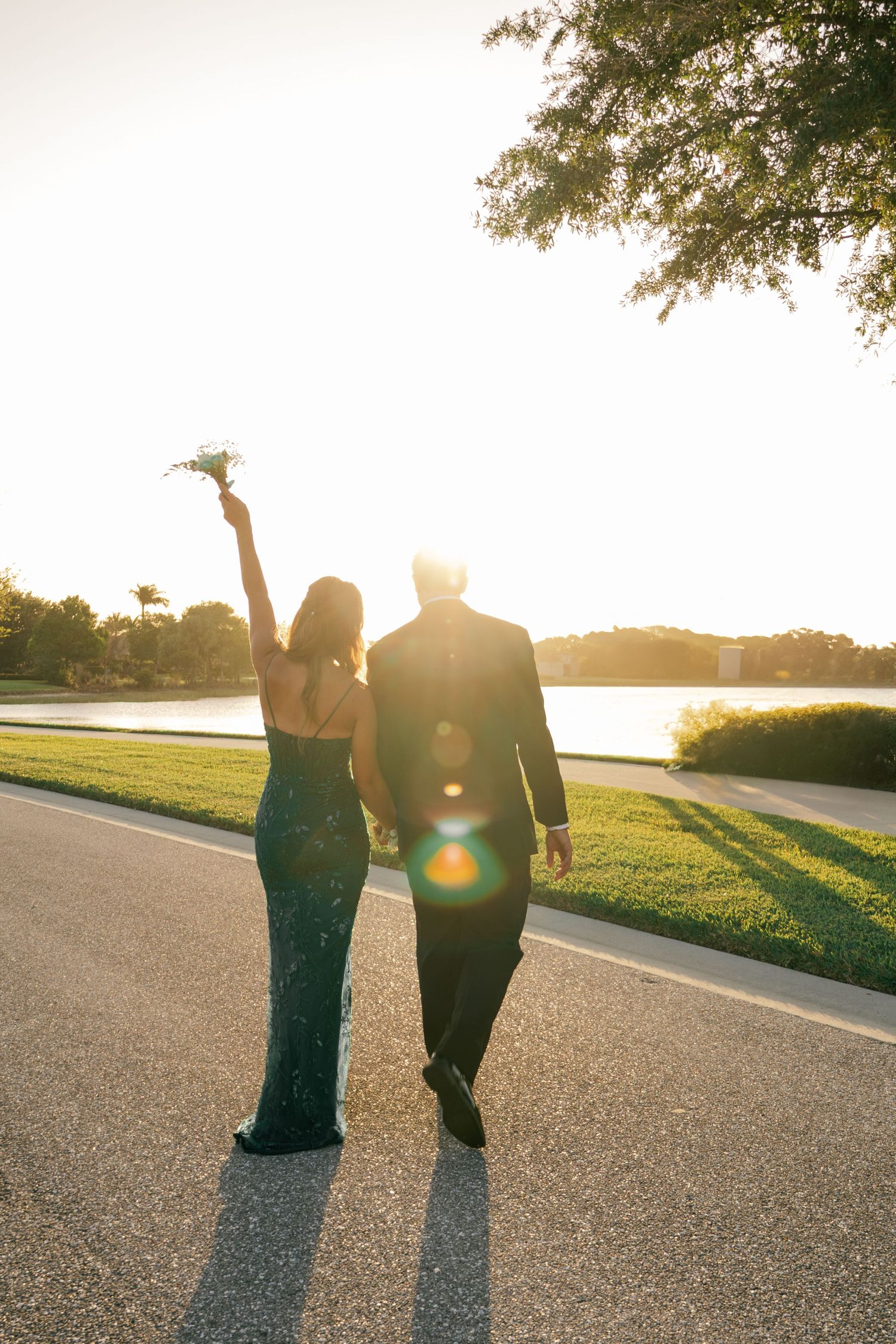 Couple in formal attire walks toward sunset lake, woman raising floral bouquet in the air.
