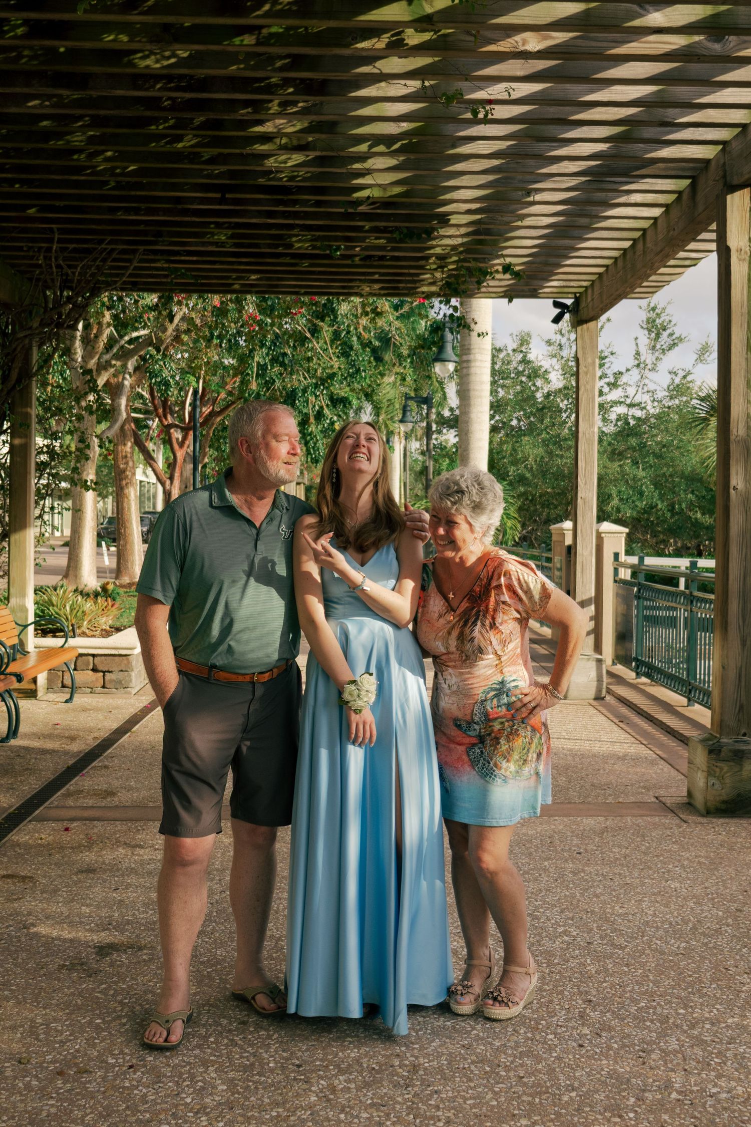 Woman in light blue gown laughs joyfully with older couple under pergola in lush outdoor setting.