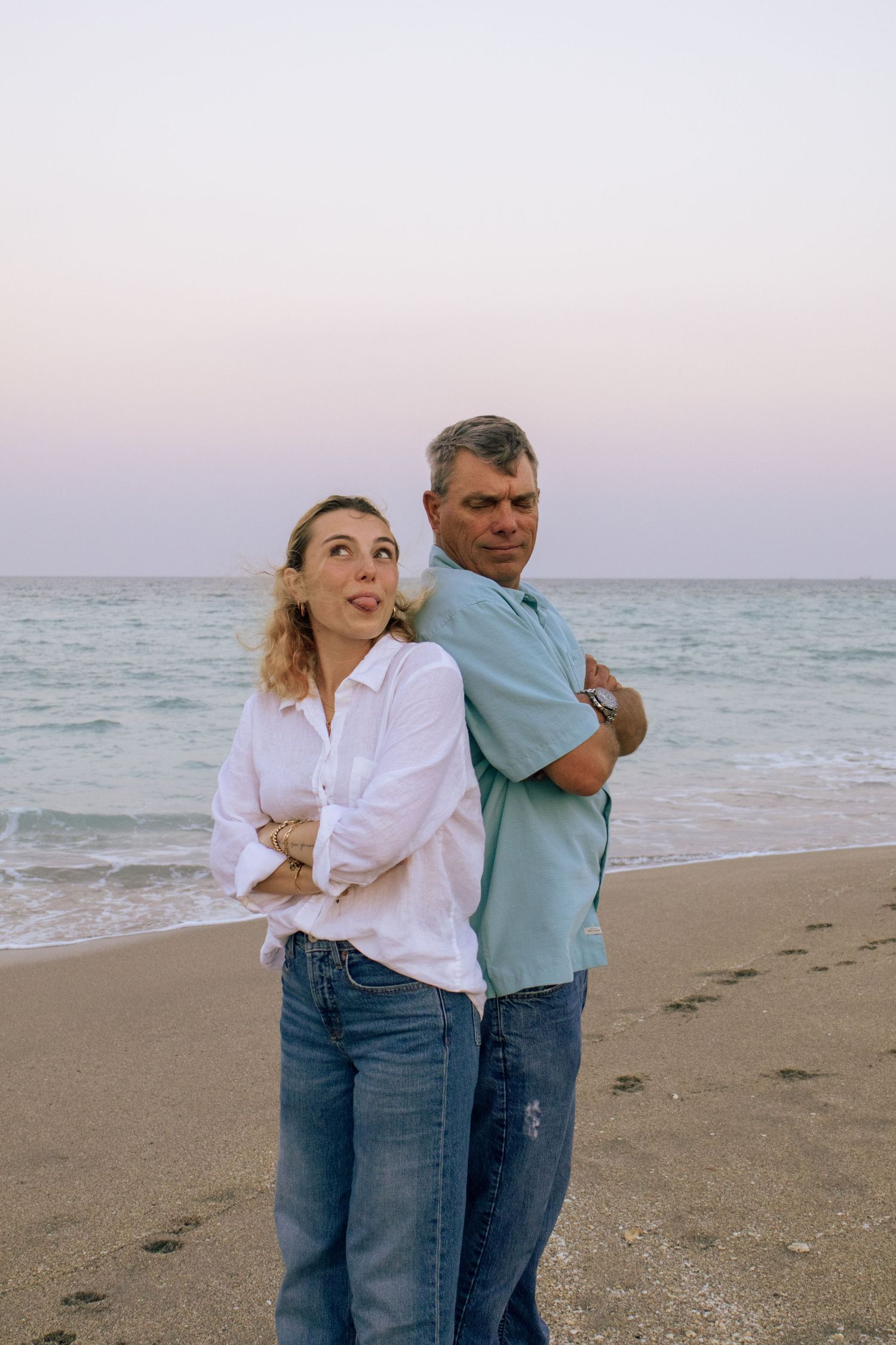 Couple standing back-to-back on a beach at dusk, arms crossed, with ocean waves and pastel sky behind them.