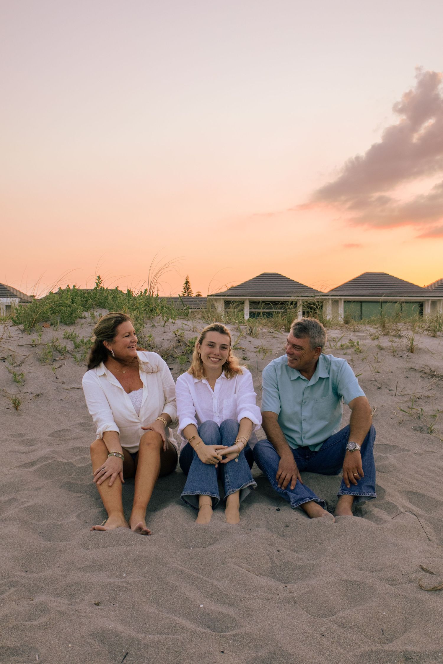 Family of three sitting on sandy beach at sunset, smiling and laughing together near coastal dunes.