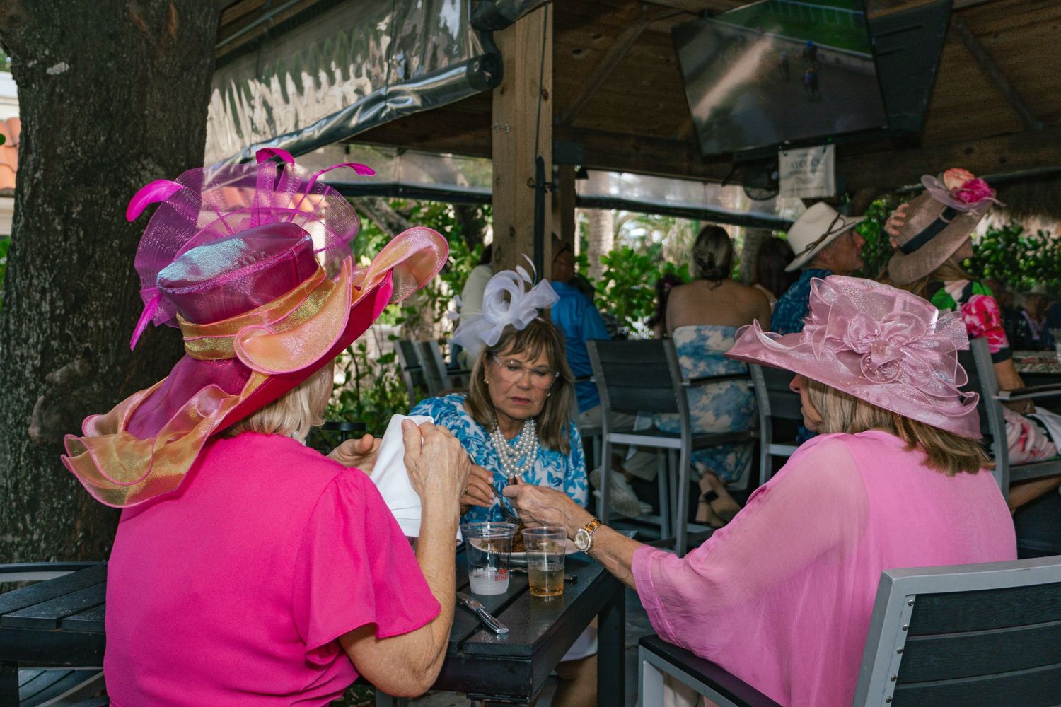 Three women wearing elegant pink fascinators and hats socialize at an outdoor Kentucky Derby party.