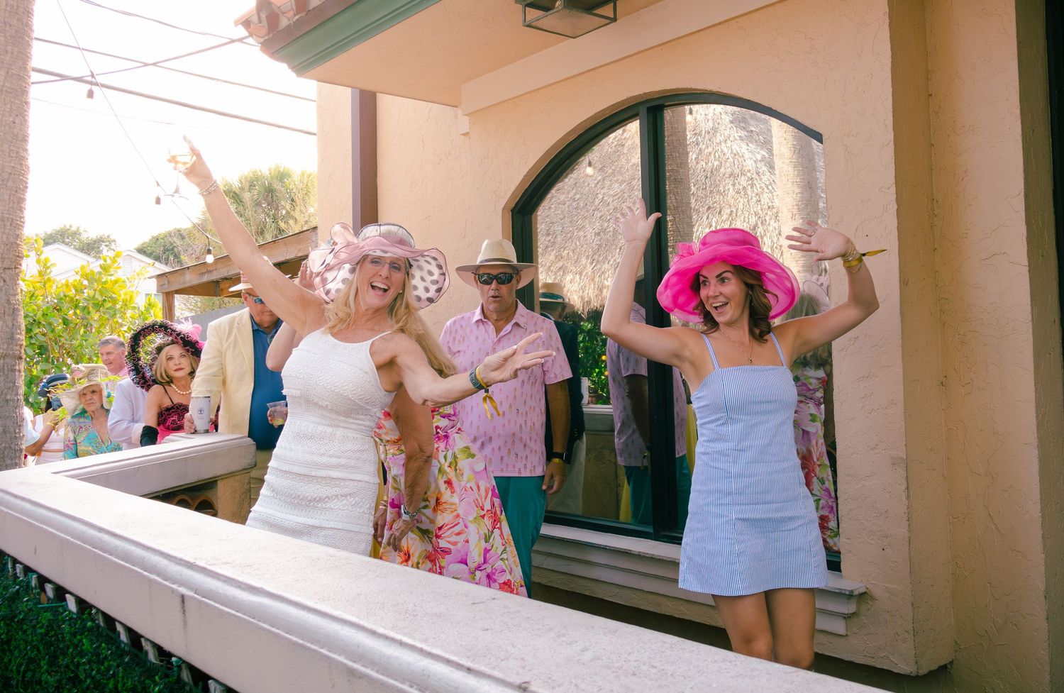 Two women in pink Derby hats celebrating with raised arms at a lively outdoor Kentucky Derby party.