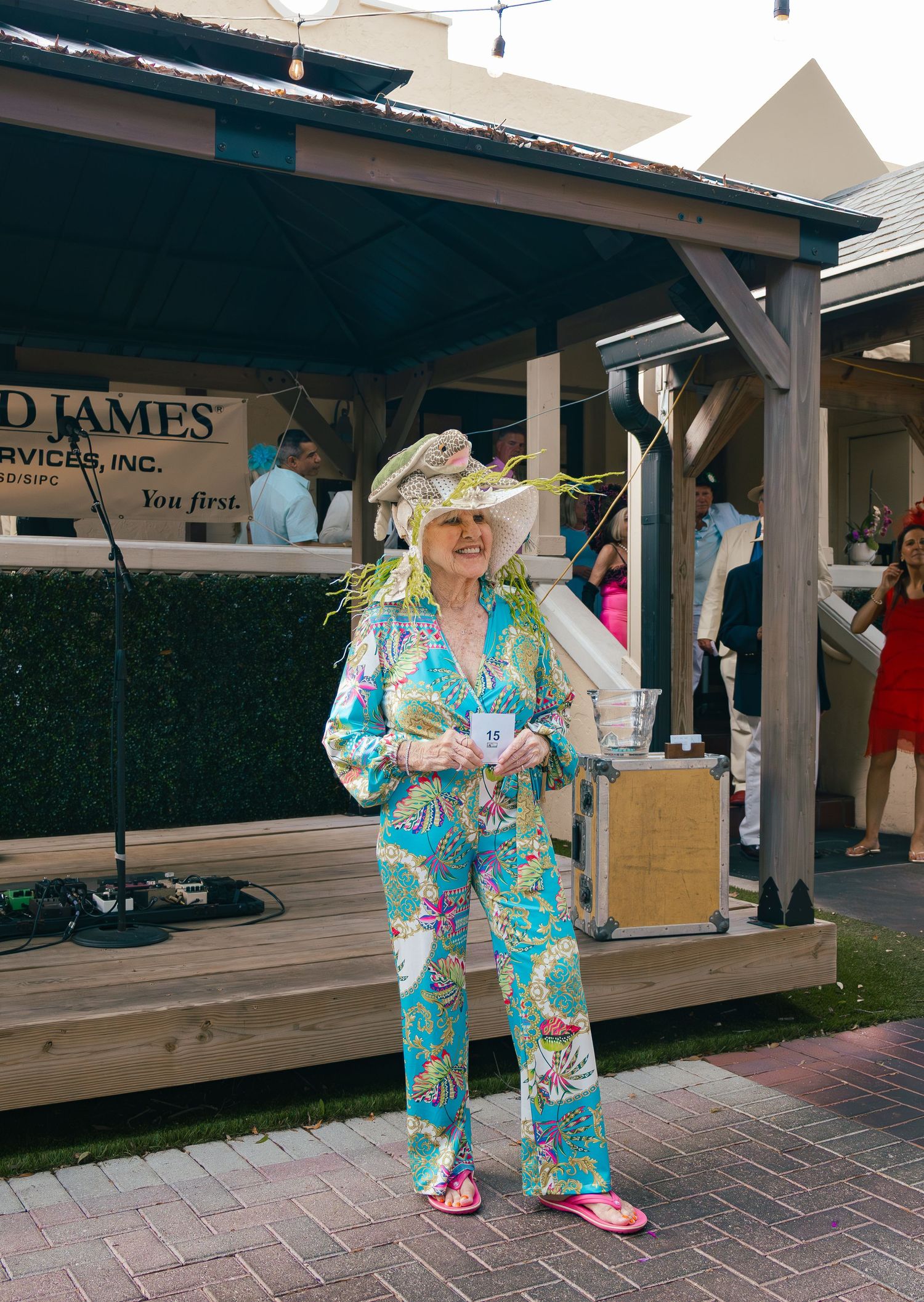 Senior woman in colorful tropical suit and elaborate floral hat at outdoor garden party.