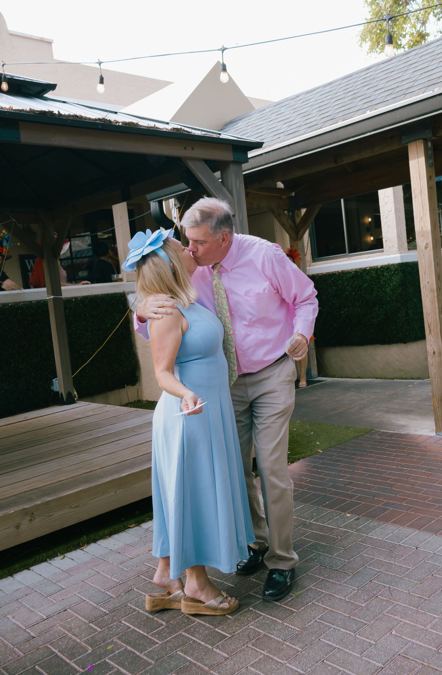 Couple sharing a kiss outdoors, woman in blue dress with fascinator hat, man in pink shirt.