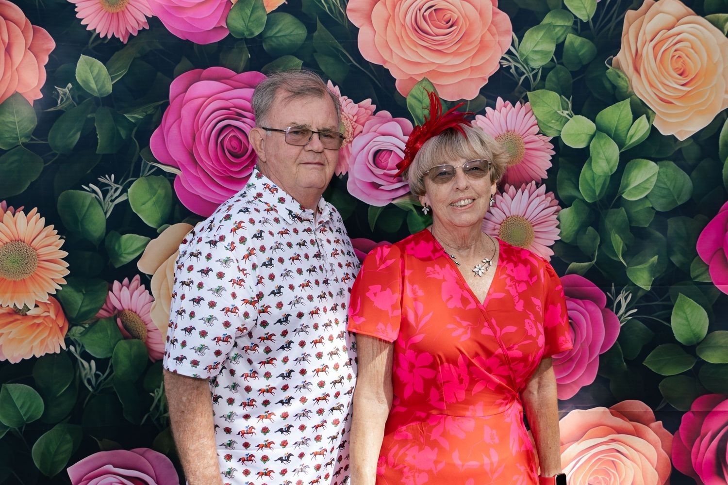 Couple dressed in festive attire posing in front of a colorful floral rose backdrop at a Derby event.