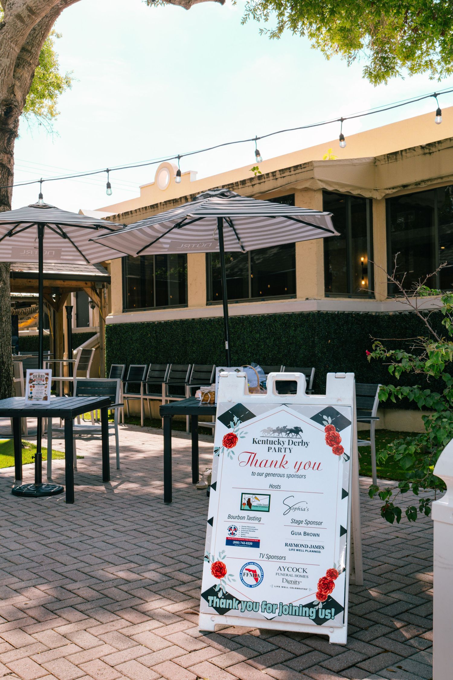 A Kentucky Derby party thank you sign on a brick patio with string lights, umbrellas, and lush greenery outside a restaurant.