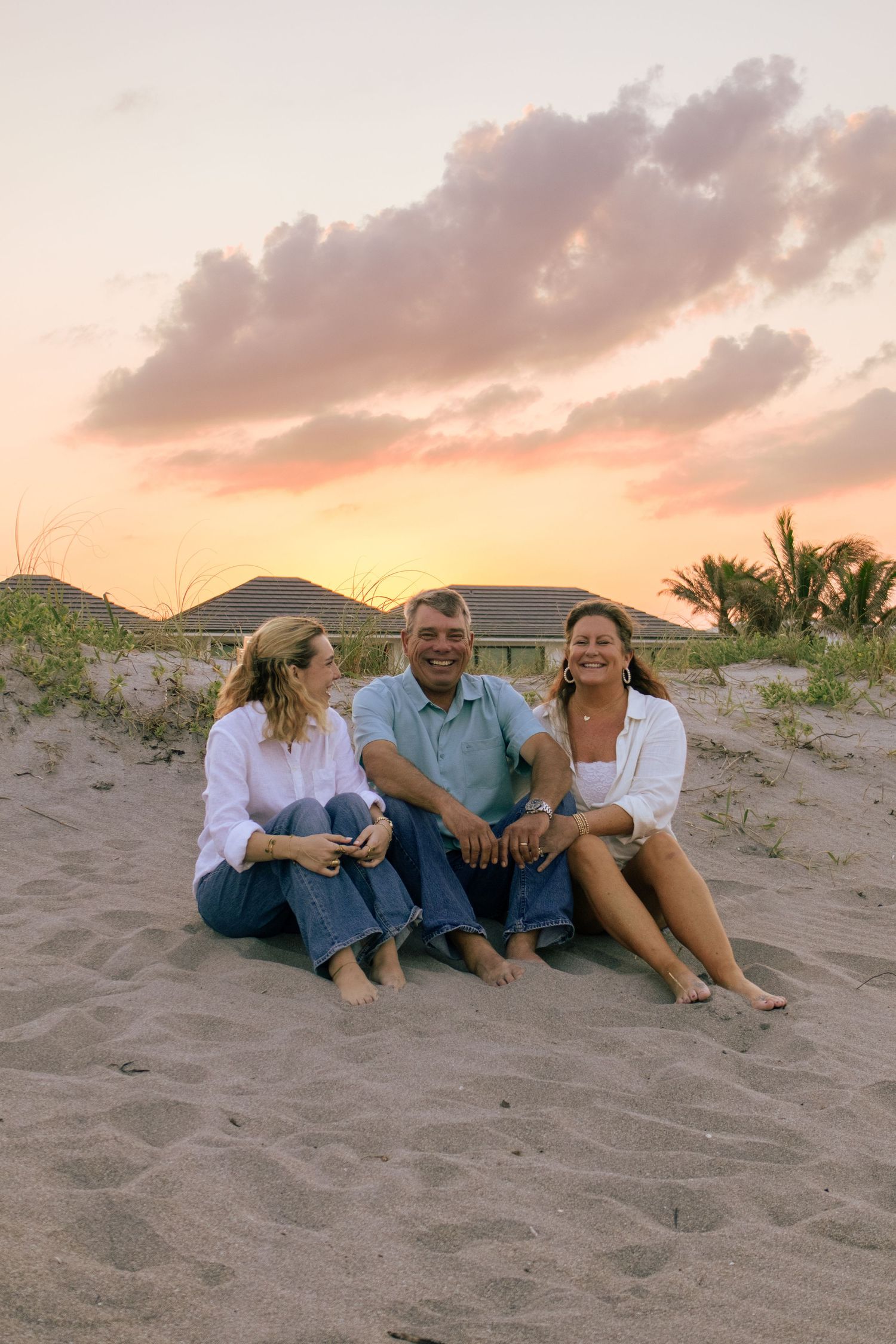 Three people sitting barefoot on sandy beach at sunset, smiling together with colorful pink sky and palm trees in background.