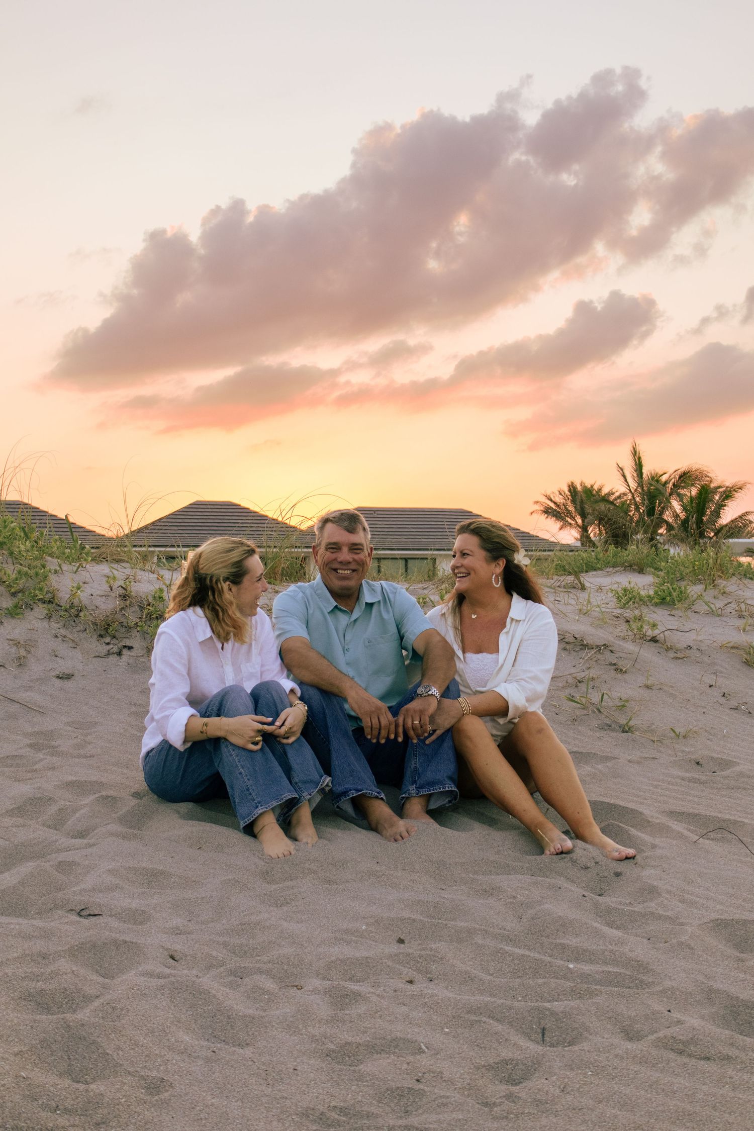 Three adults sitting barefoot on sandy beach at sunset, smiling and enjoying each others company near coastal dunes.