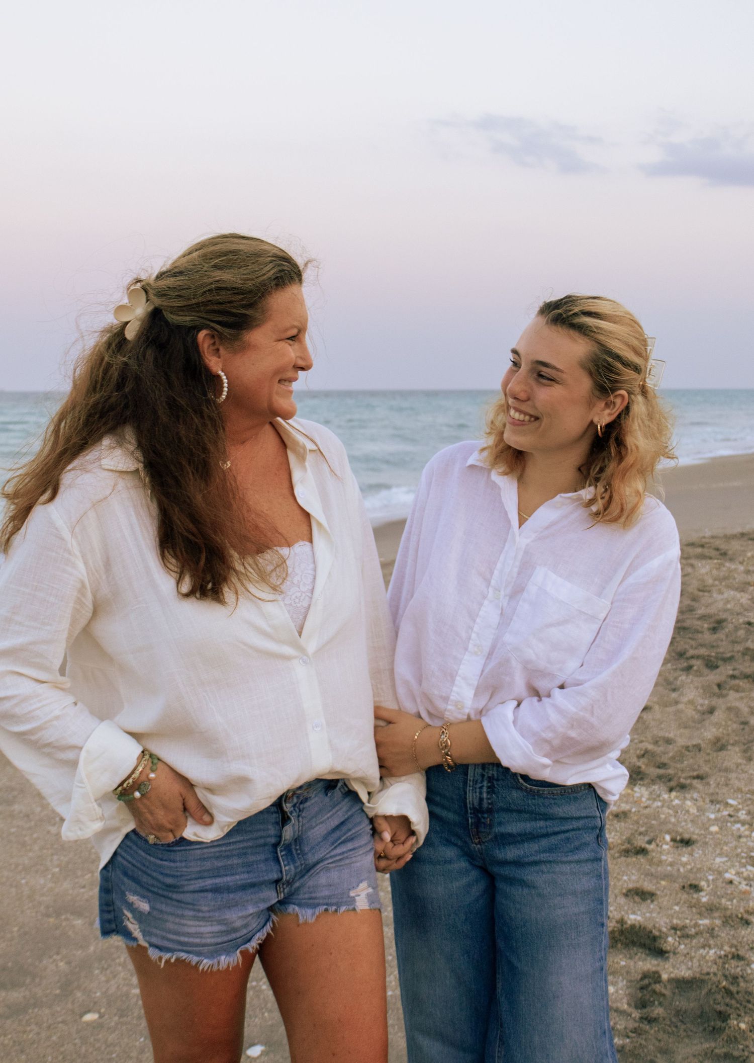 Two women in white shirts smiling at each other on beach at dusk near ocean waves.