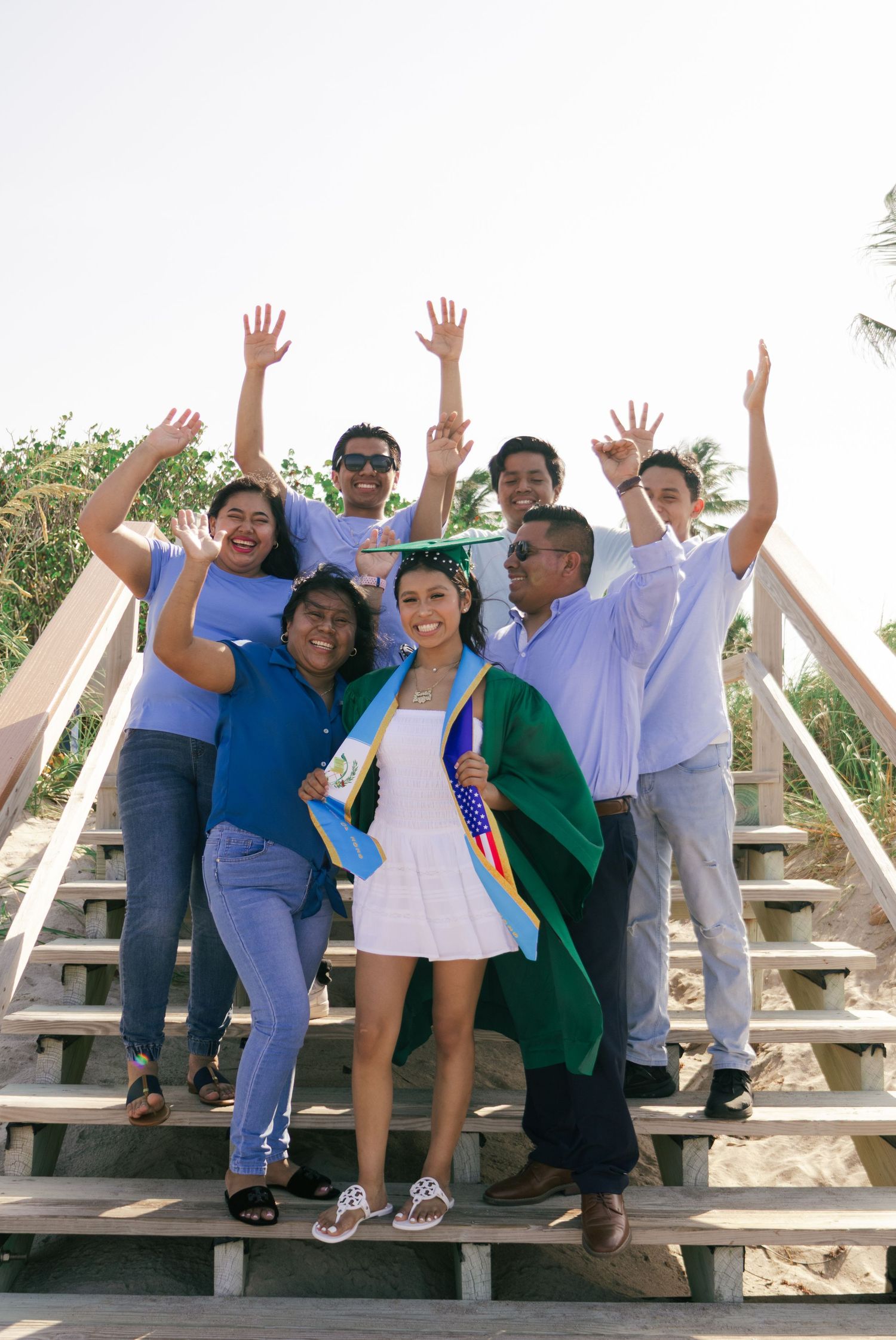 Group of seven people celebrating on wooden stairs at beach, holding flags, arms raised in joy.