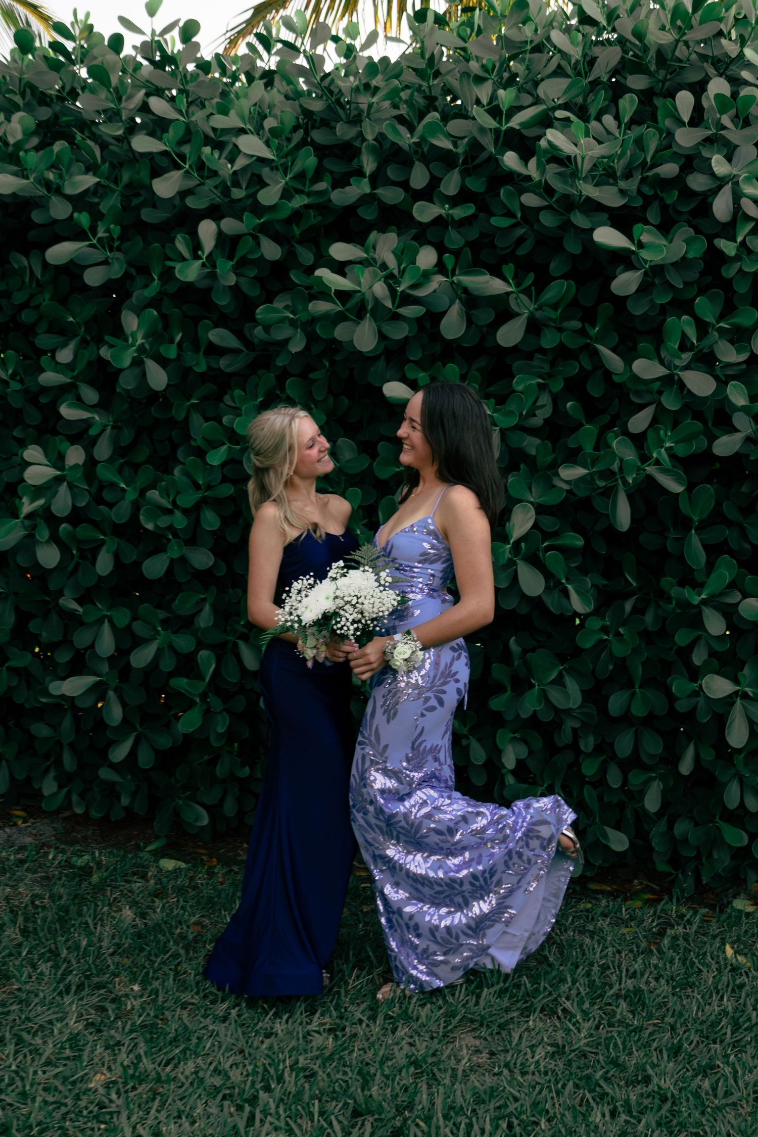 Two women in blue formal dresses laugh together in front of lush green hedge wall.