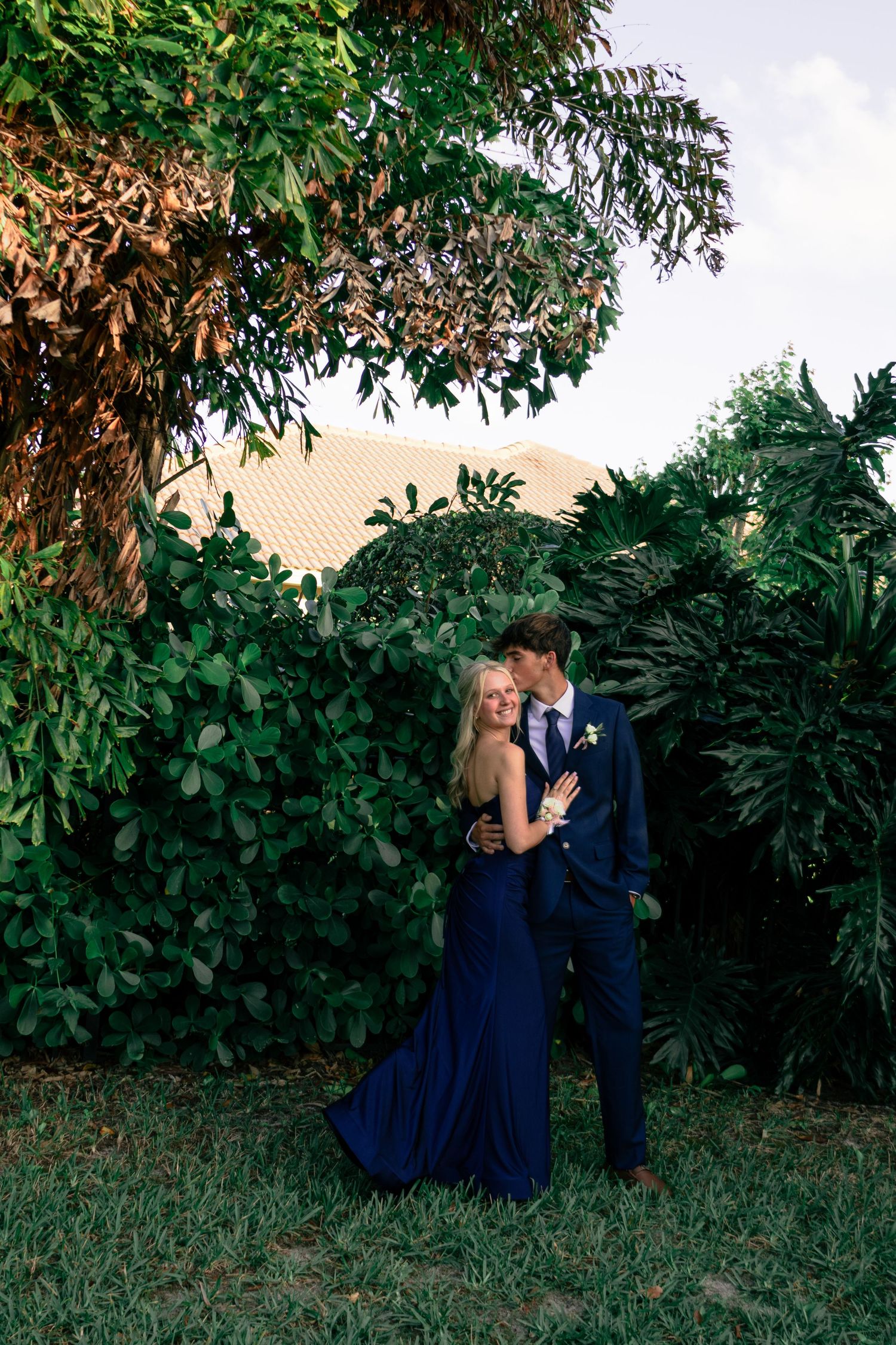Elegant couple posing in a tropical garden, dressed in matching navy blue prom outfits.