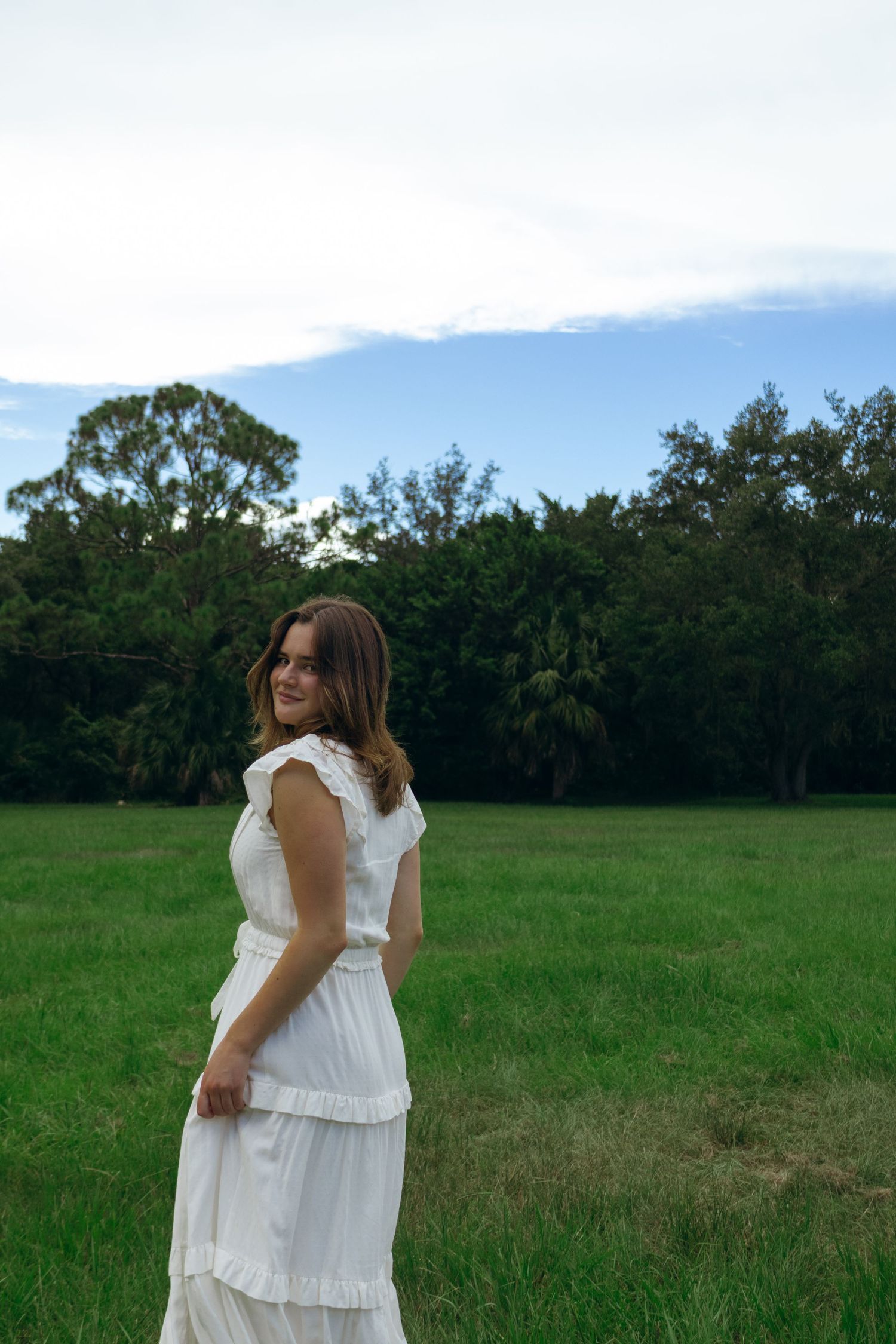 Young woman in white ruffled dress standing in green field, smiling over her shoulder at camera.