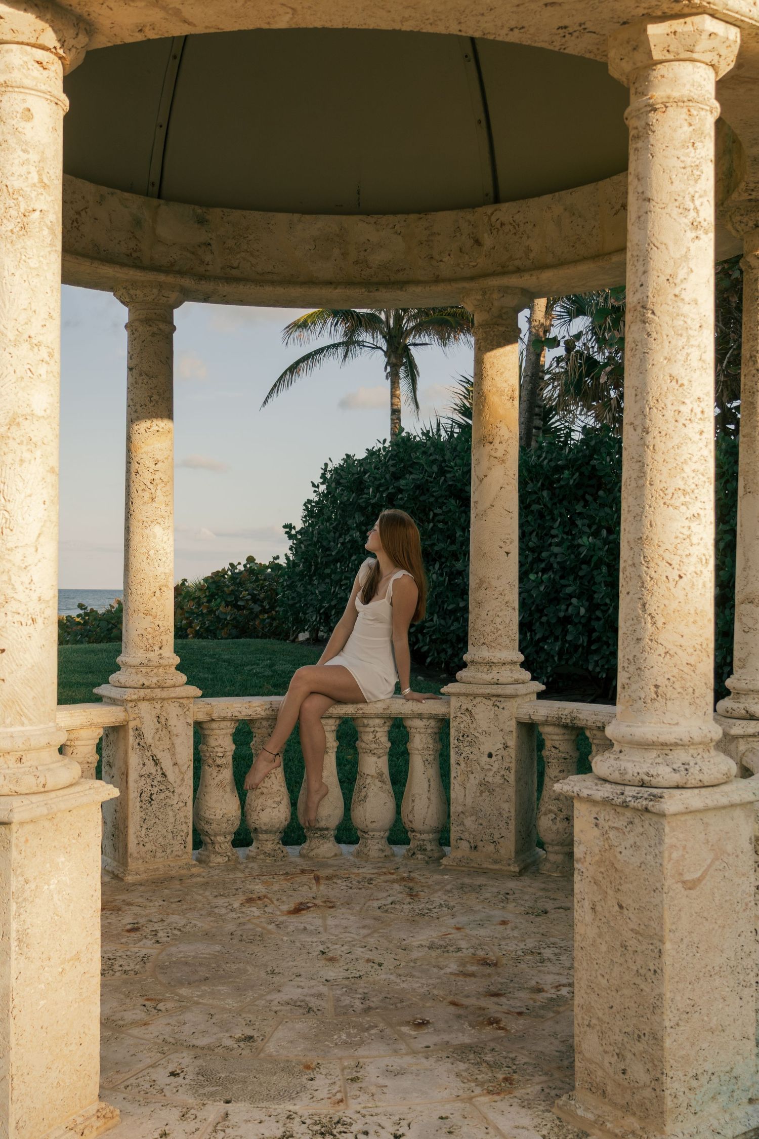 Young woman in white dress sitting on stone balustrade in elegant gazebo overlooking tropical ocean landscape.