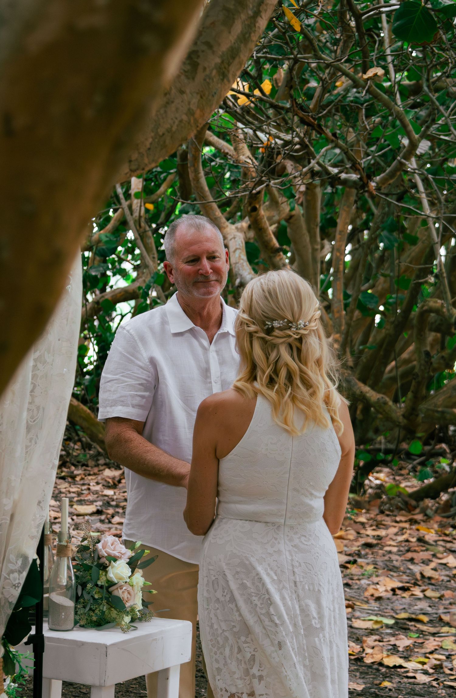 Groom smiling warmly at his bride during an outdoor wedding ceremony beneath a canopy of tropical trees.