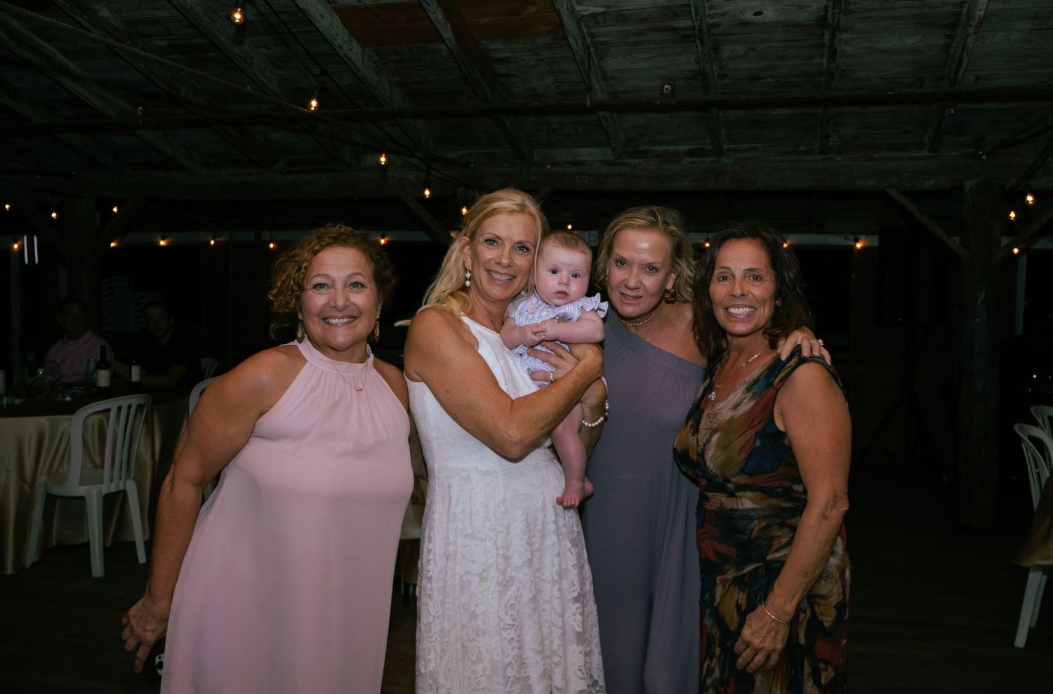 Bride holding baby surrounded by three smiling female wedding guests at reception.