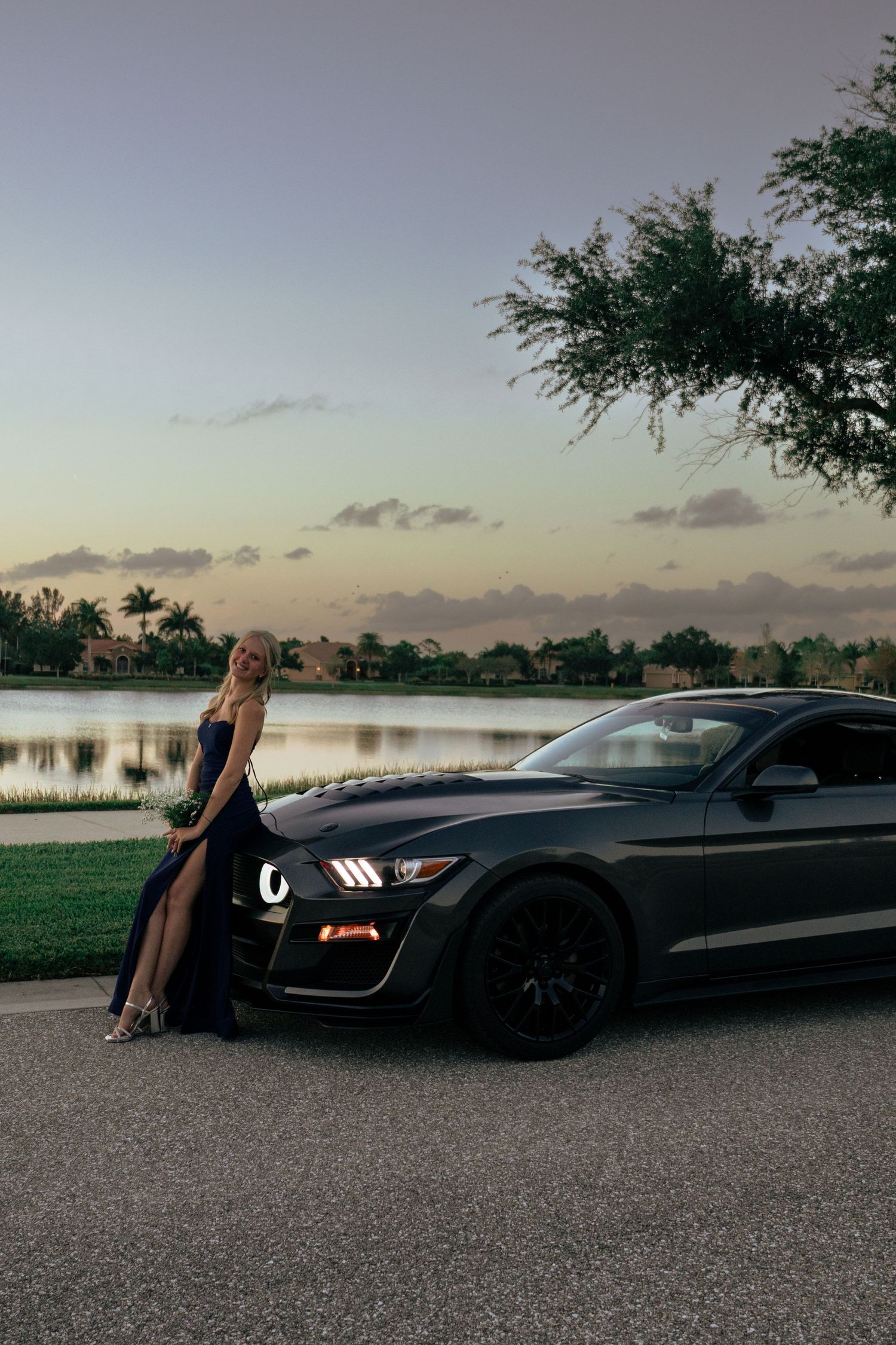 Elegant young woman in a black formal gown posing beside a dark Ford Mustang at dusk lakeside.