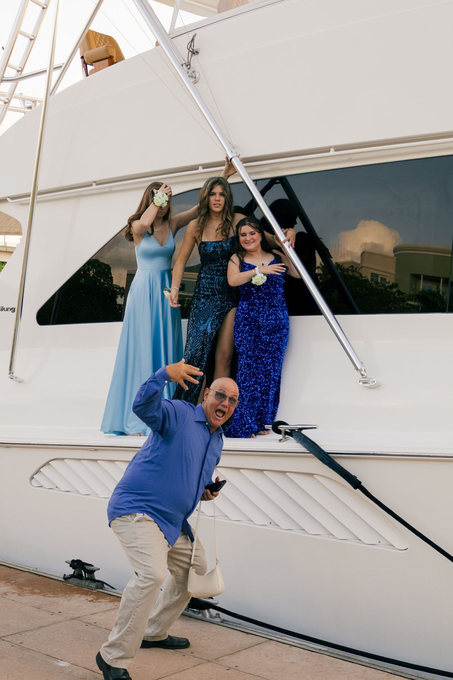 Three young women in blue prom dresses posing on a yacht with a man in foreground.