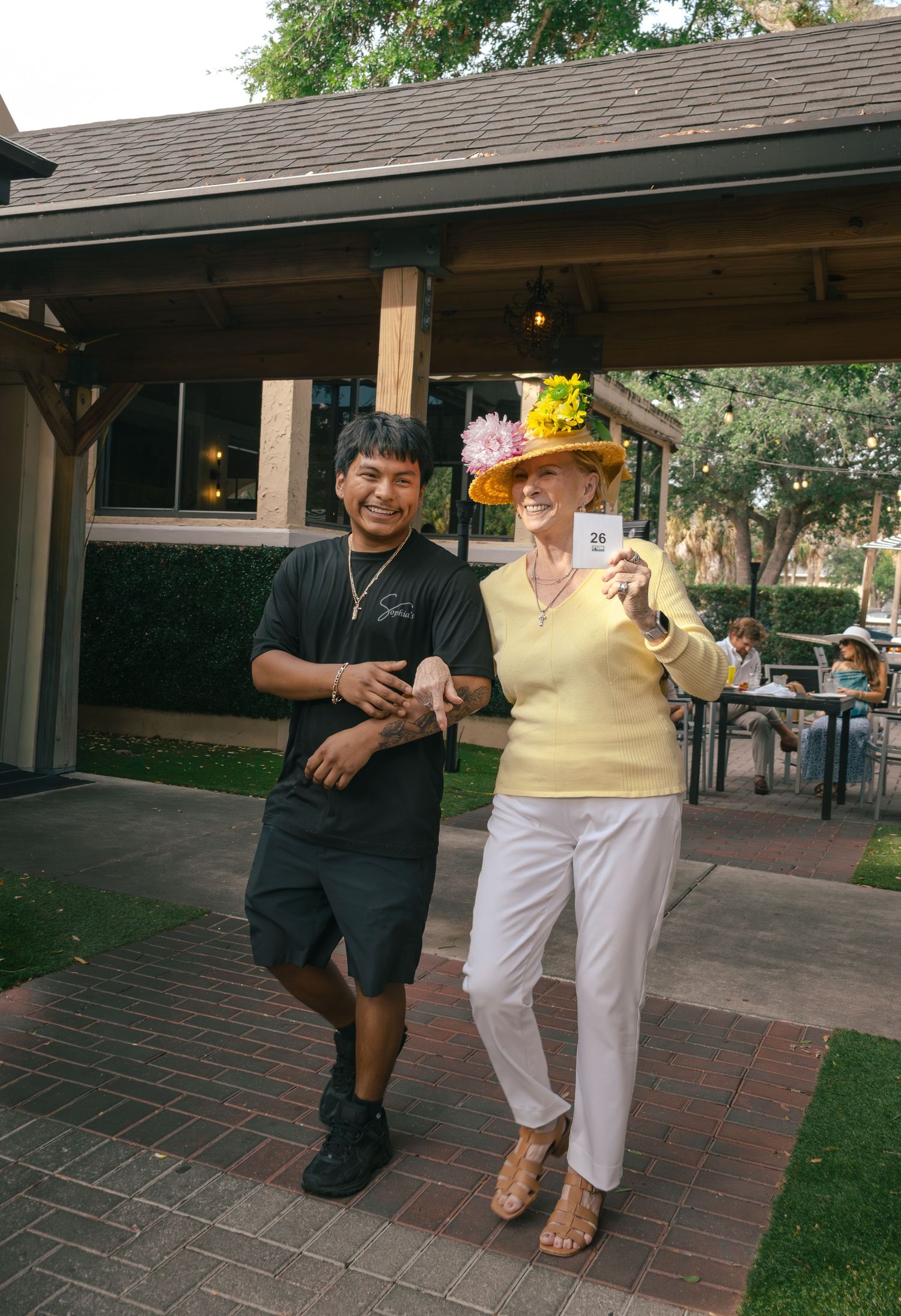 Man and woman posing outdoors, she wears a yellow floral hat and holds a card at a garden event.
