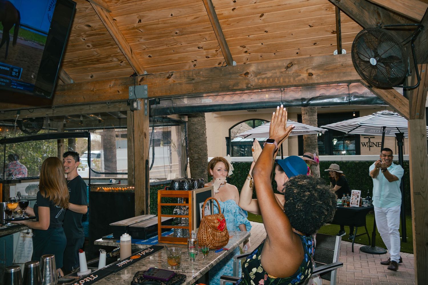 Lively bar scene with guests celebrating and raising hands at an outdoor venue with wooden ceiling.