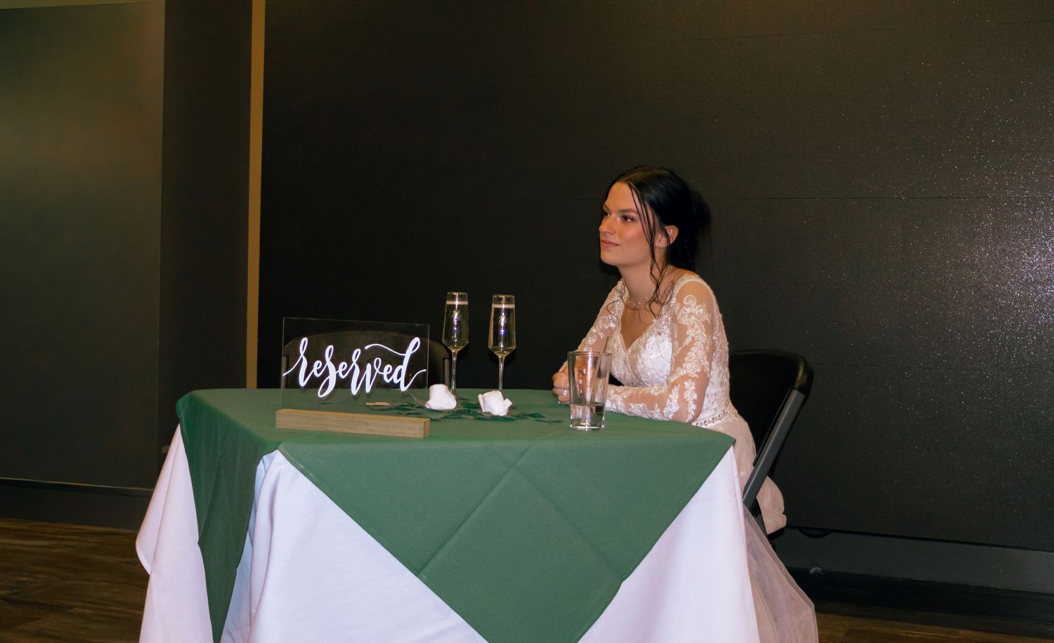 Bride in long-sleeve lace wedding gown sits alone at a green and white sweetheart table with a reserved acrylic sign.